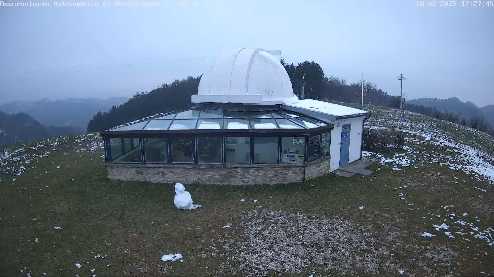 A small observatory with a glass sunroom and a dome sits on a snow-dusted hill overlooking a hazy mountain range under an overcast sky.