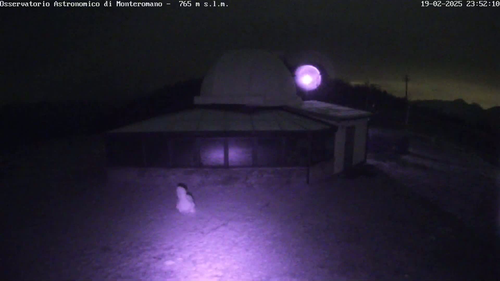 A low-light nighttime image shows a building with a dome, possibly an observatory, sitting in a snow-covered area under a dark, cloudy sky.