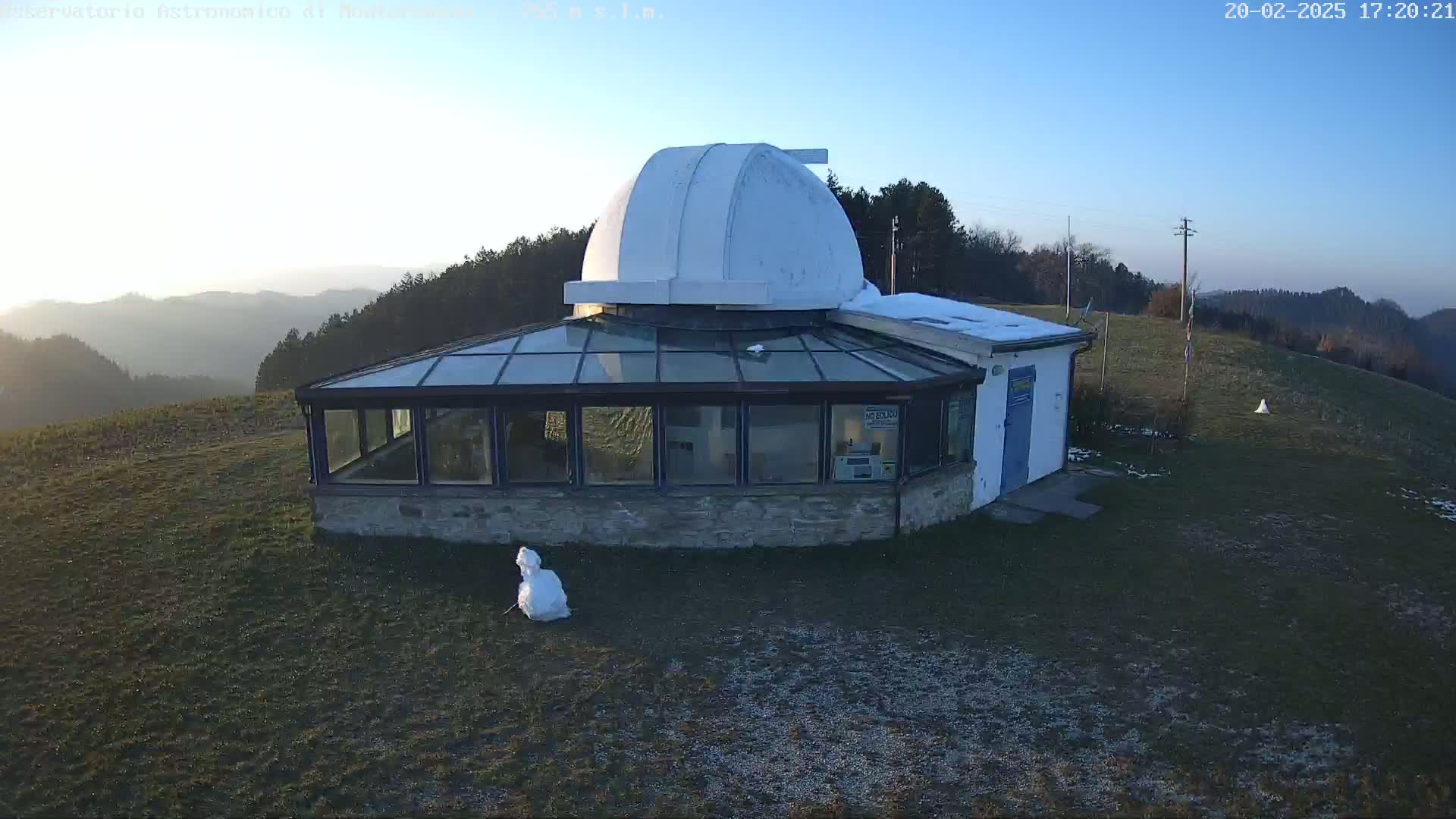 A stone building with a glass enclosure and a white dome on top sits on a grassy hill overlooking a hazy mountain range under a clear blue sky.