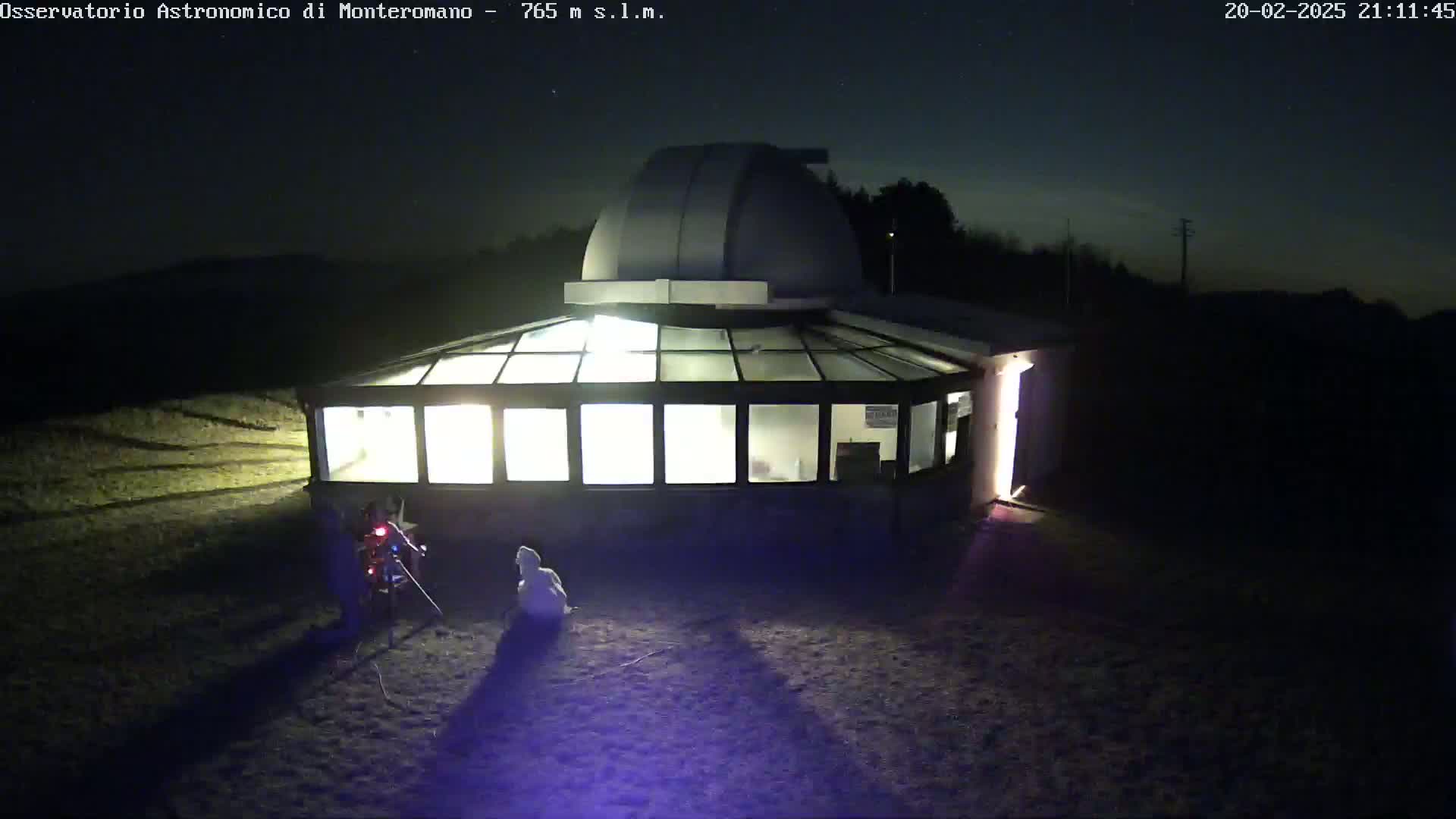 An illuminated observatory building with a dome on top is surrounded by dark, sparsely vegetated grounds under a clear night sky.