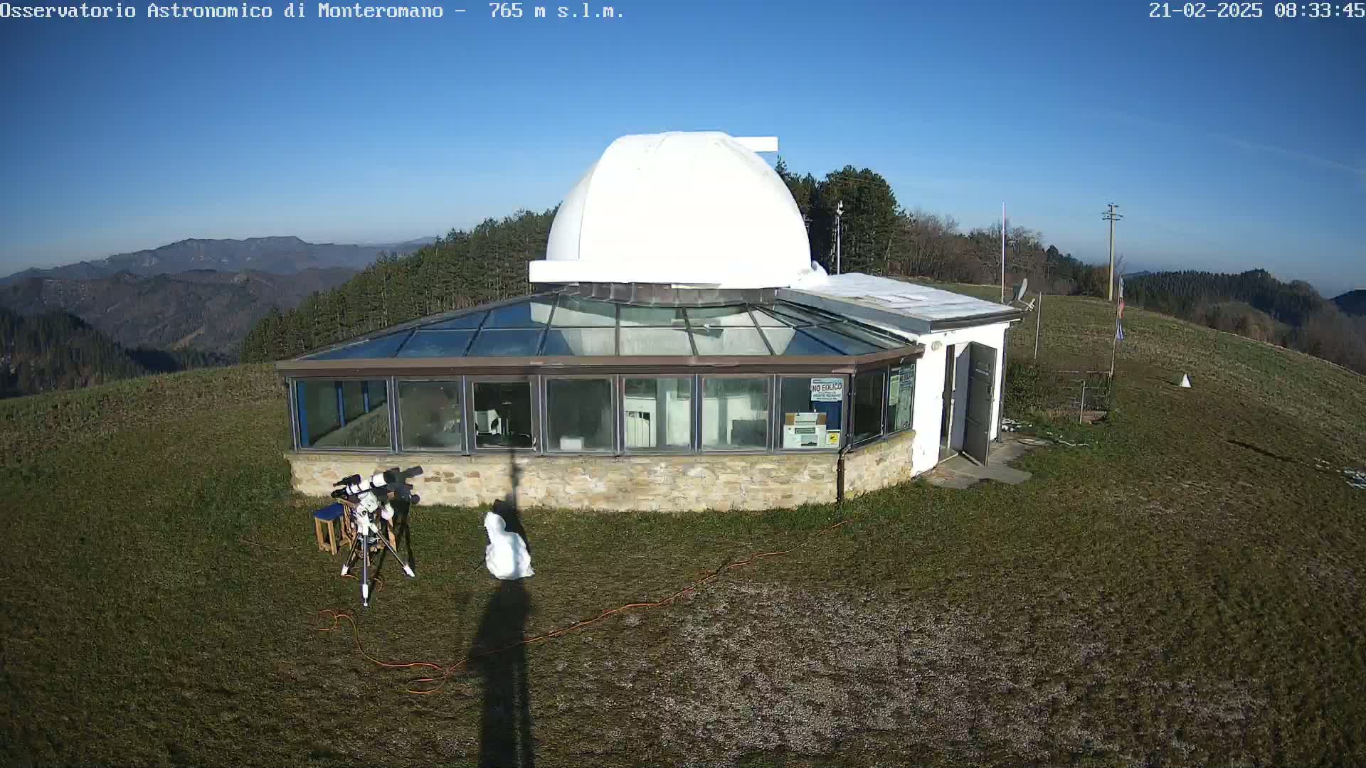 An observatory with a white dome and a glass-walled building sits on a grassy hilltop under a clear blue sky.