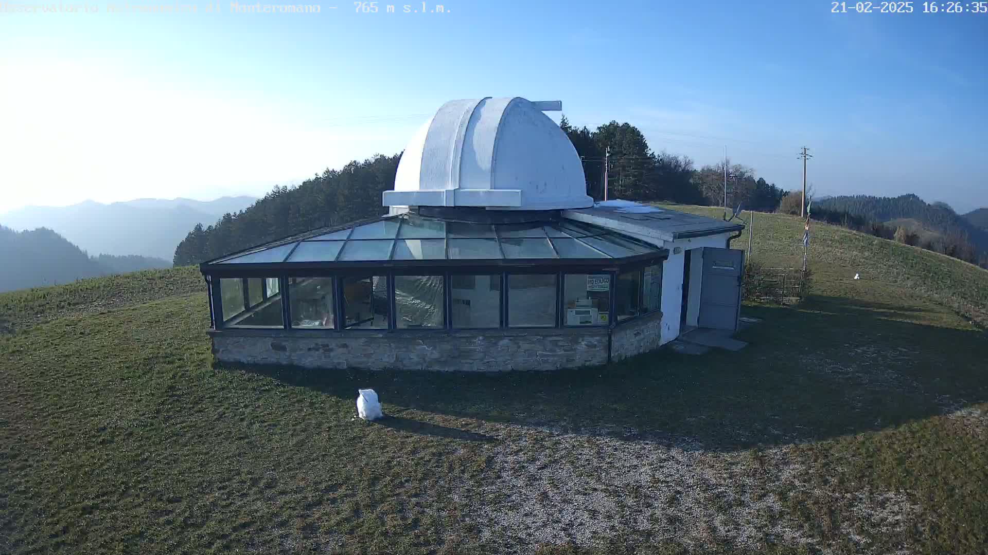 A white observatory dome sits atop a stone building with a glass sunroom, overlooking a grassy hill and distant, hazy mountains under a clear blue sky.