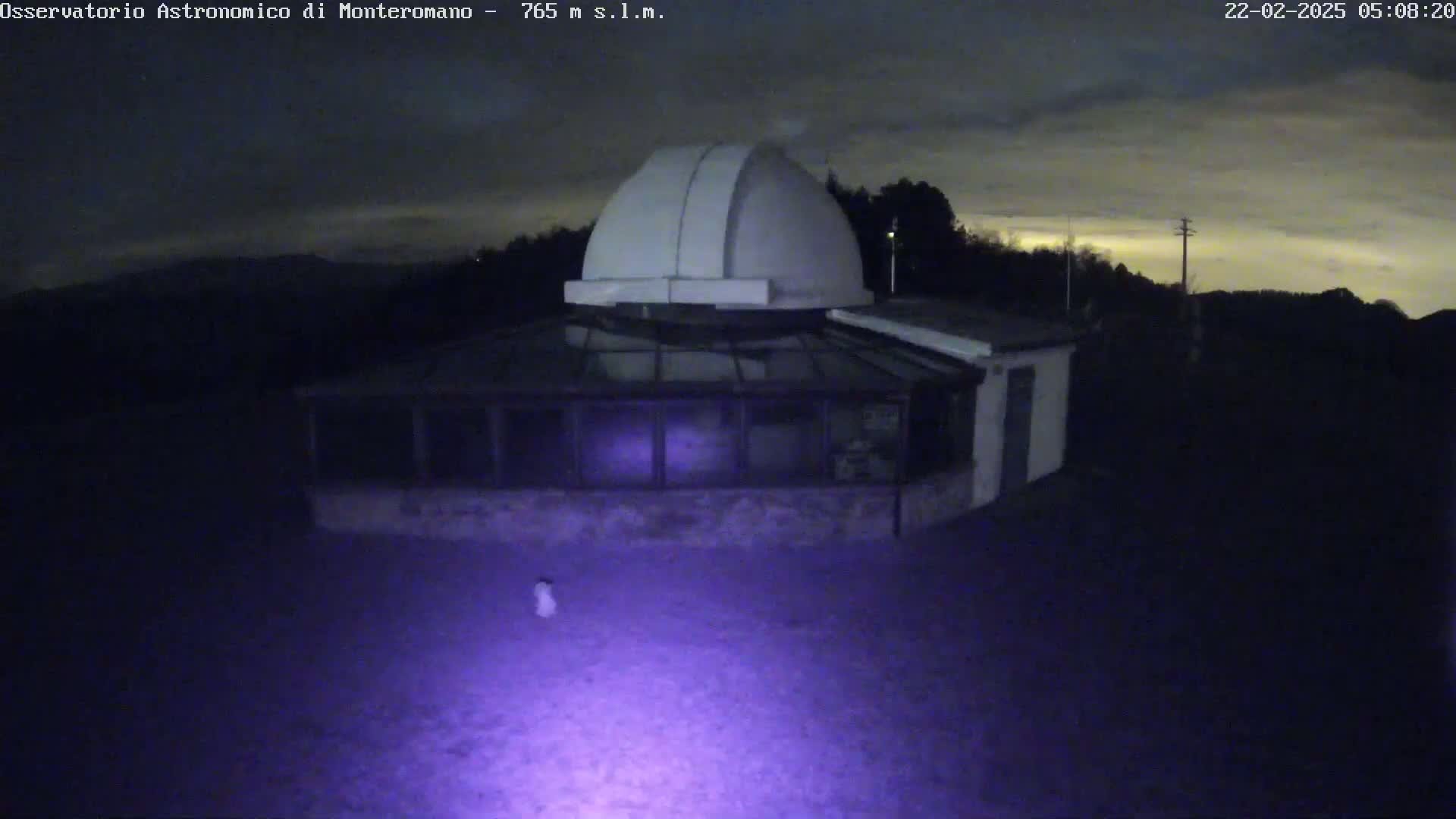 A white observatory dome sits atop a building with glass walls, illuminated by a purple light on a dark, possibly cloudy night.