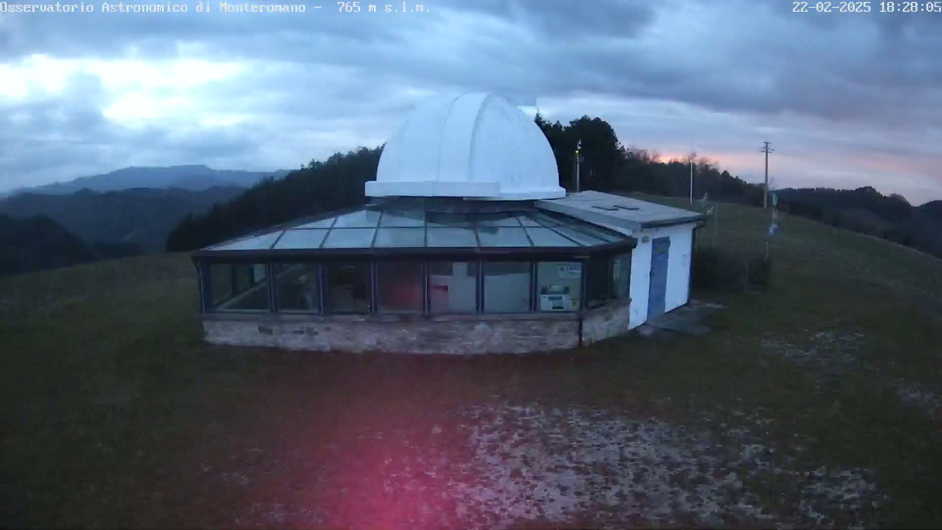 A white-domed observatory building with a glass-roofed addition sits on a hilltop overlooking a mountainous landscape under a cloudy, twilight sky.