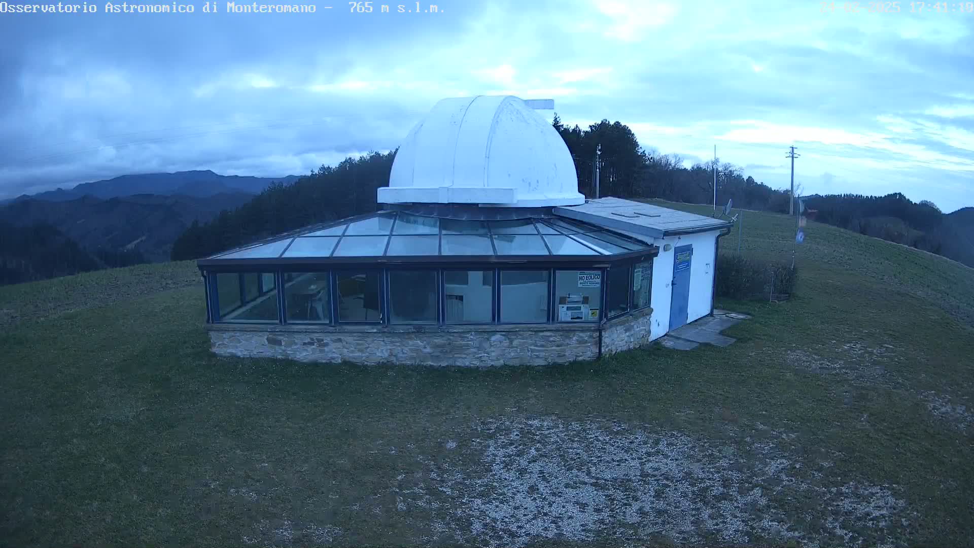 A stone building with a glass conservatory and a white dome sits on a grassy hill overlooking a mountain range under a cloudy sky.