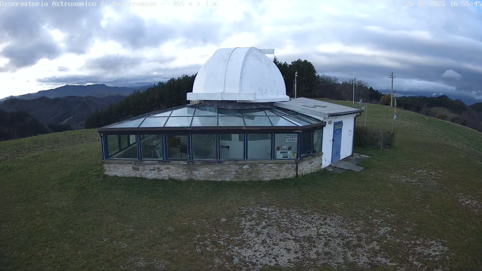 An observatory with a white dome sits atop a grassy hill under a cloudy sky, overlooking a distant mountain range.