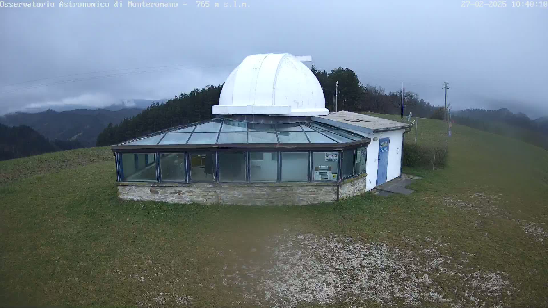 A small observatory building with a glass enclosure and a white dome sits atop a grassy hill under an overcast sky.