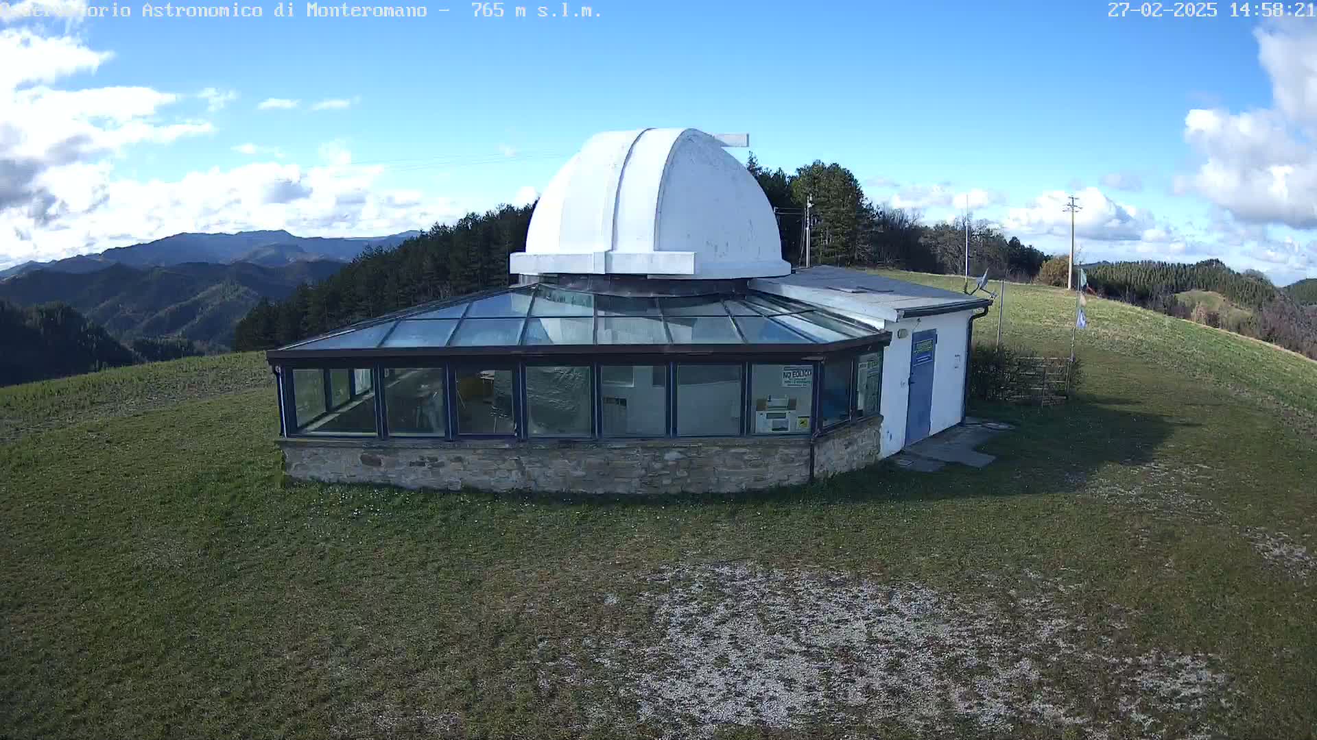 An observatory with a white dome sits atop a grassy hill under a partly cloudy, sunny sky, overlooking a mountain range.
