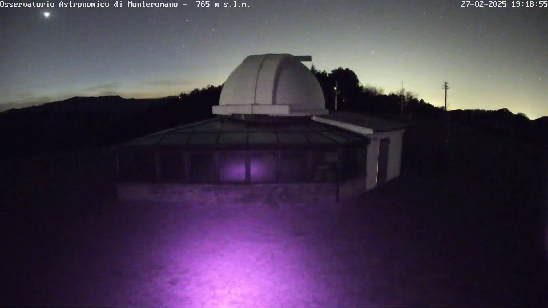 A white dome-shaped observatory building with a glass-roofed addition is illuminated by a purple light at night under a clear, starry sky.