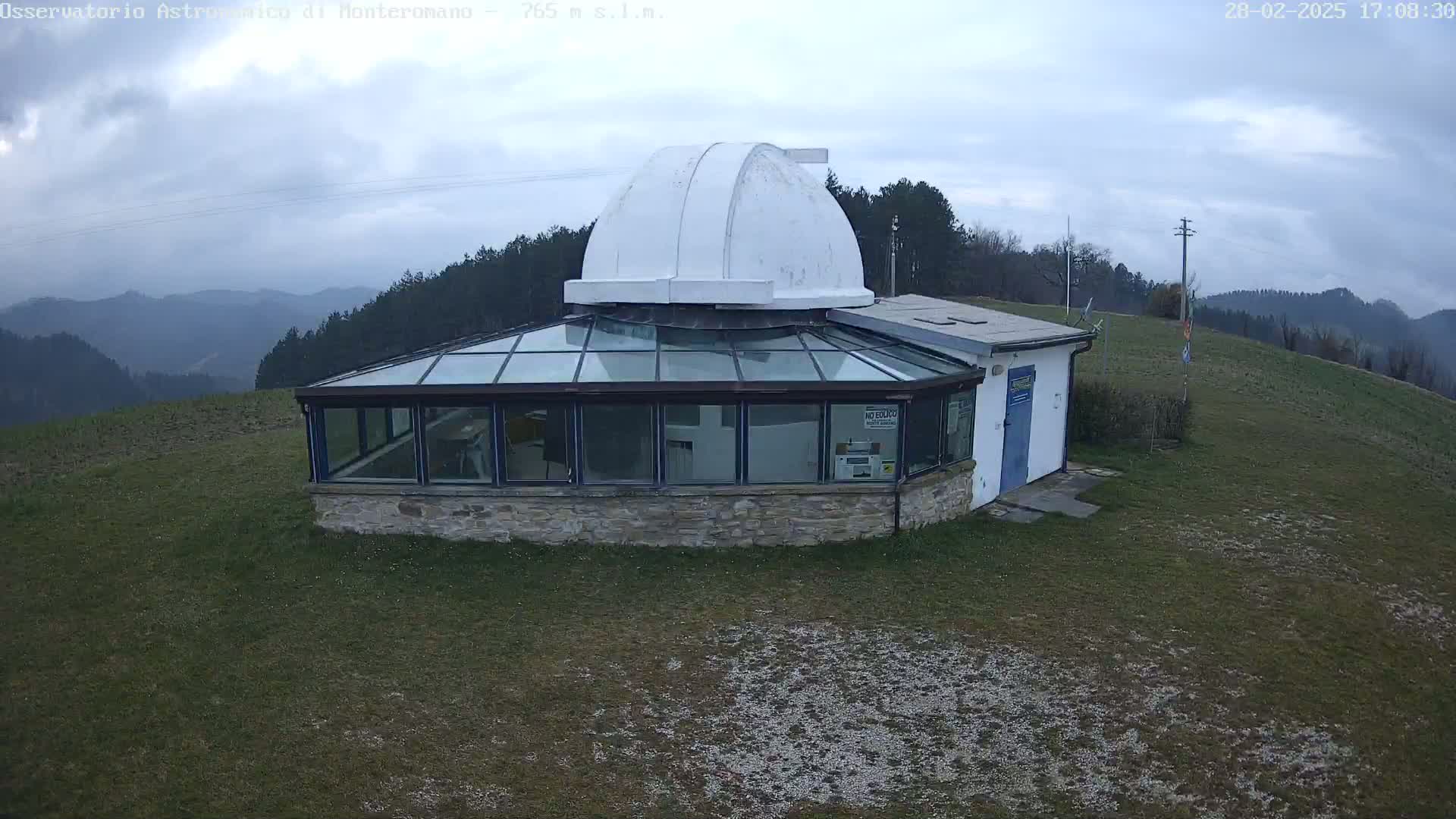 A stone building with a glass conservatory and a white dome sits on a grassy hilltop overlooking a hazy mountain range under a cloudy sky.