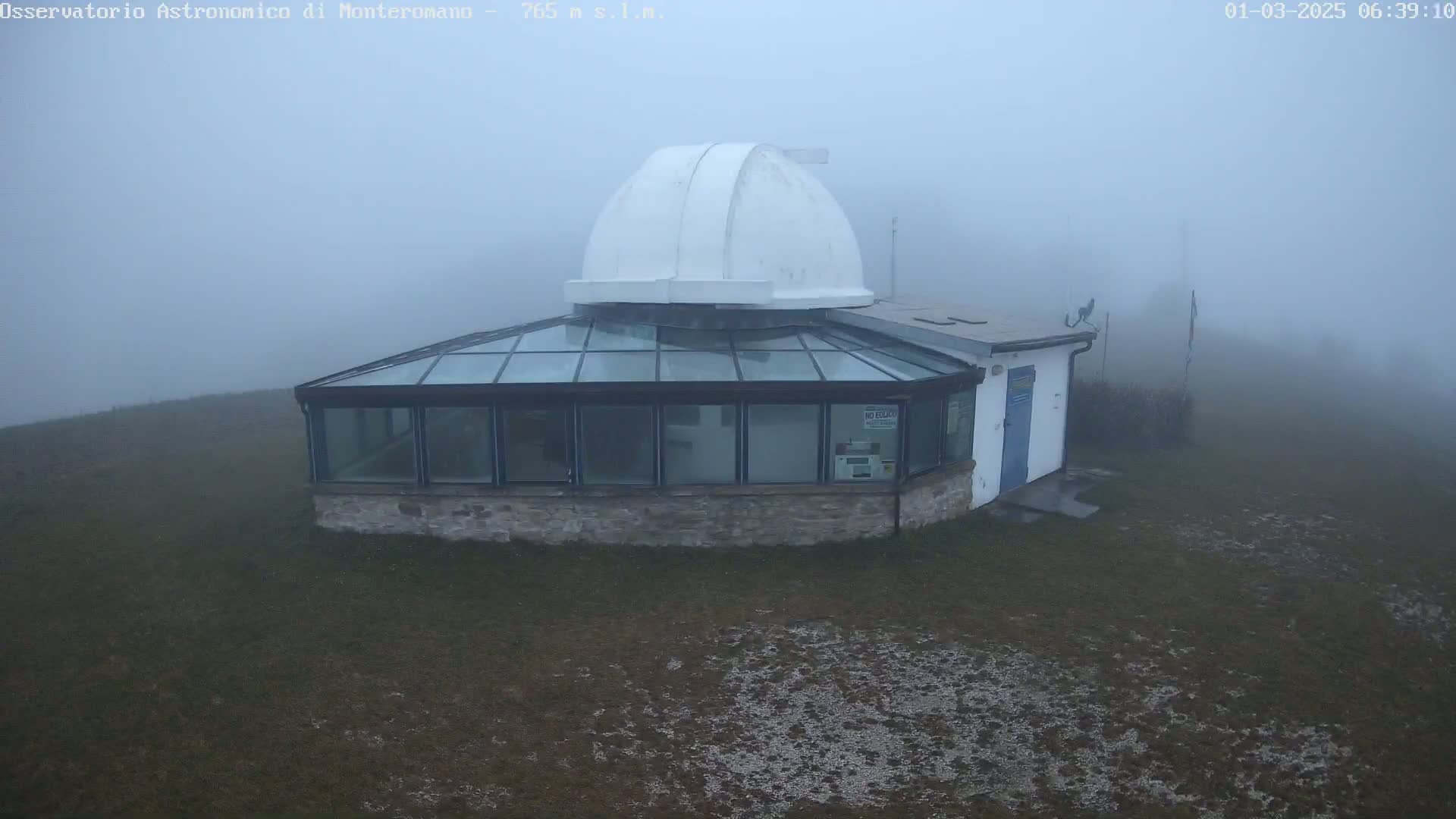 A stone observatory building with a glass-roofed addition and a white dome sits in heavy fog on a grassy area.