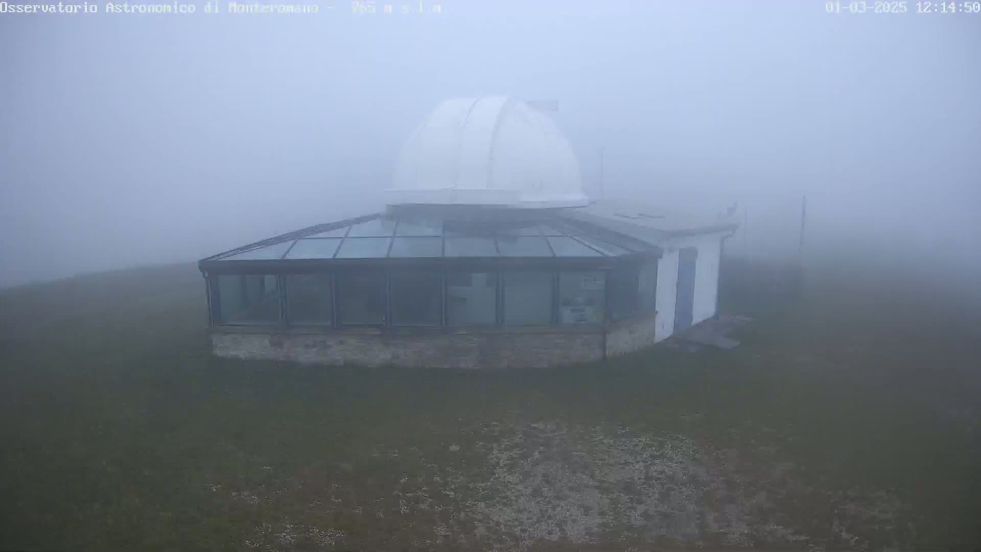 A building with a glass roof and a white dome sits on a grassy area in thick fog.