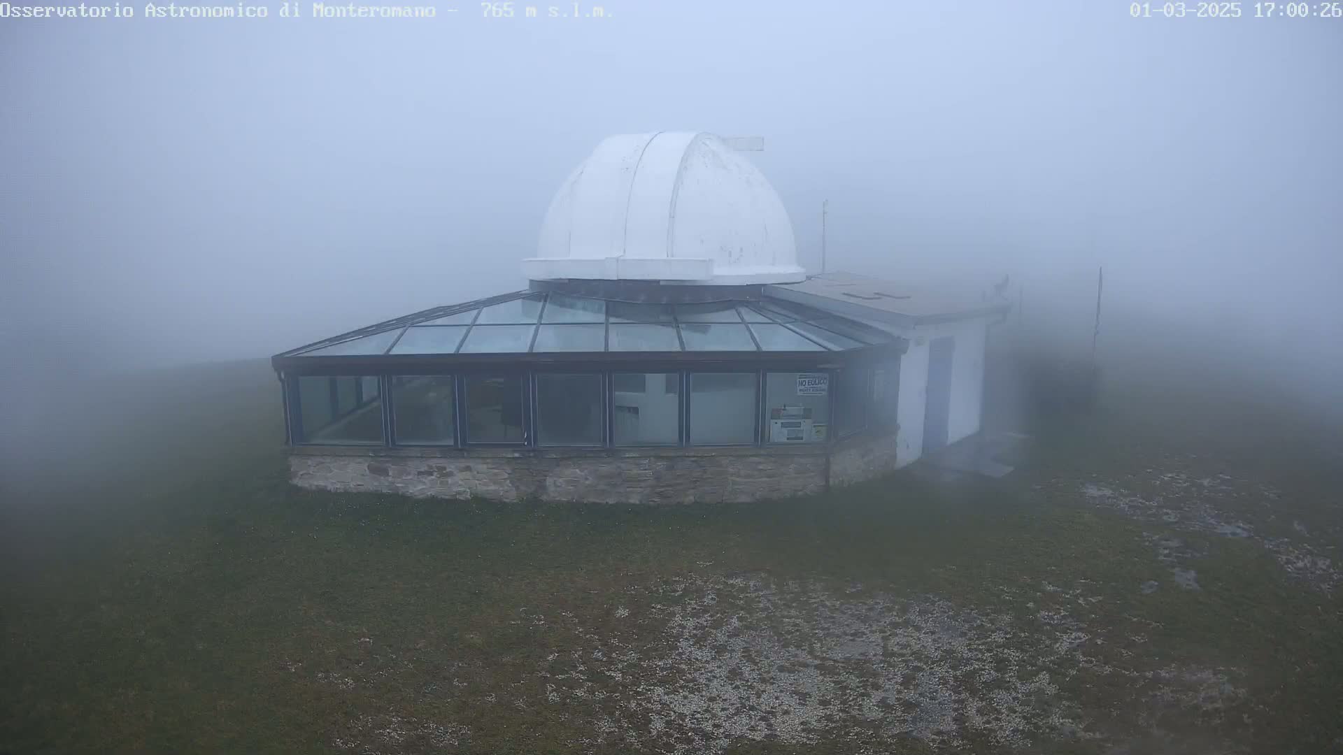 A small observatory with a dome and glass walls sits on a grassy area shrouded in thick fog.