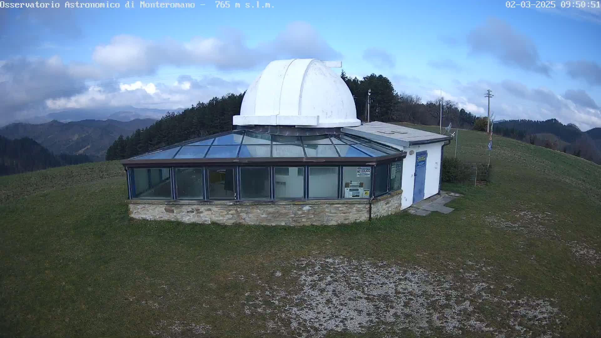 A white-domed observatory building with a glass-roofed addition sits atop a grassy hill under a partly cloudy blue sky.