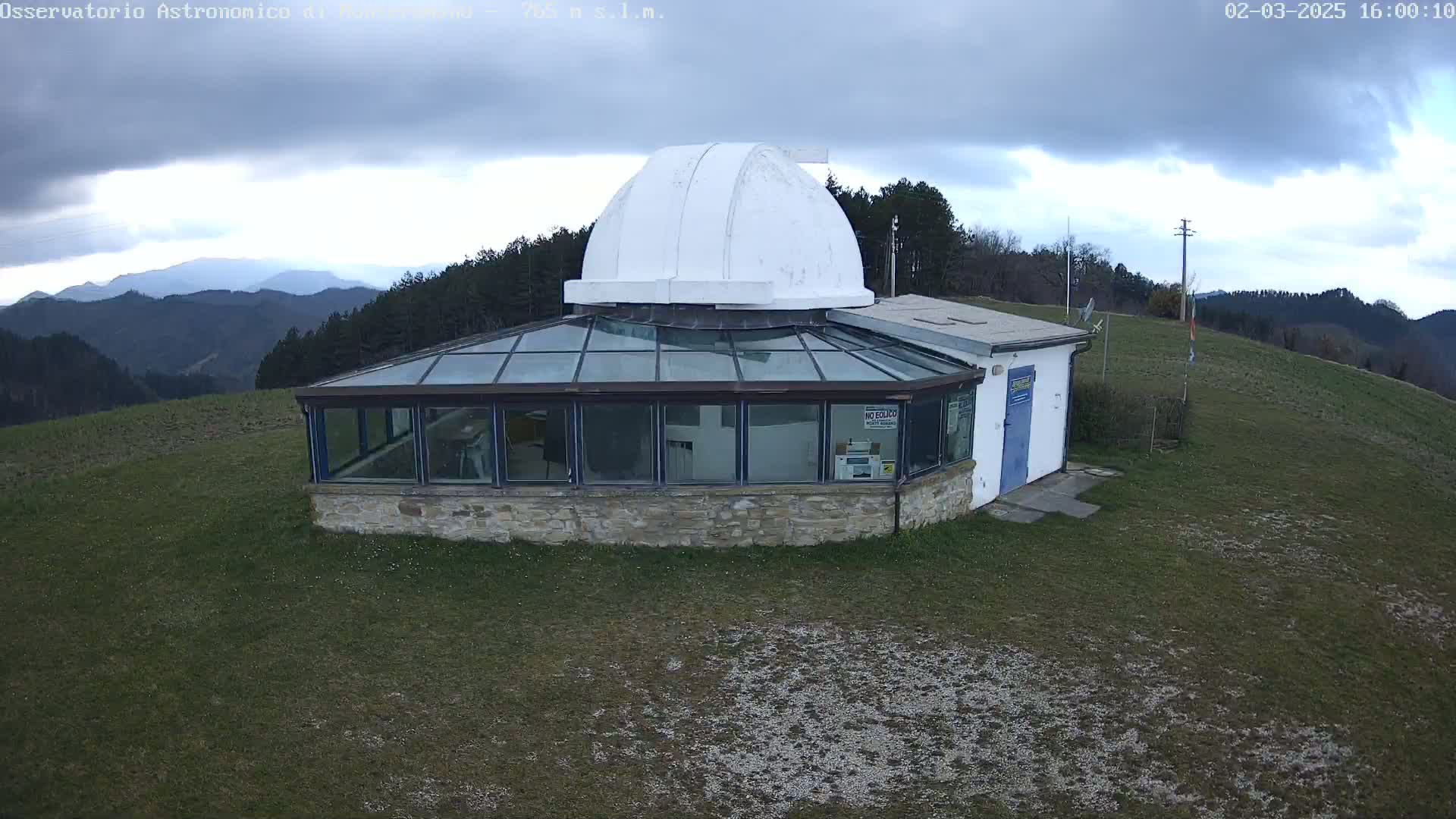 A stone building with a glass sunroom and a white dome sits atop a grassy hill under a cloudy sky, overlooking distant mountains.
