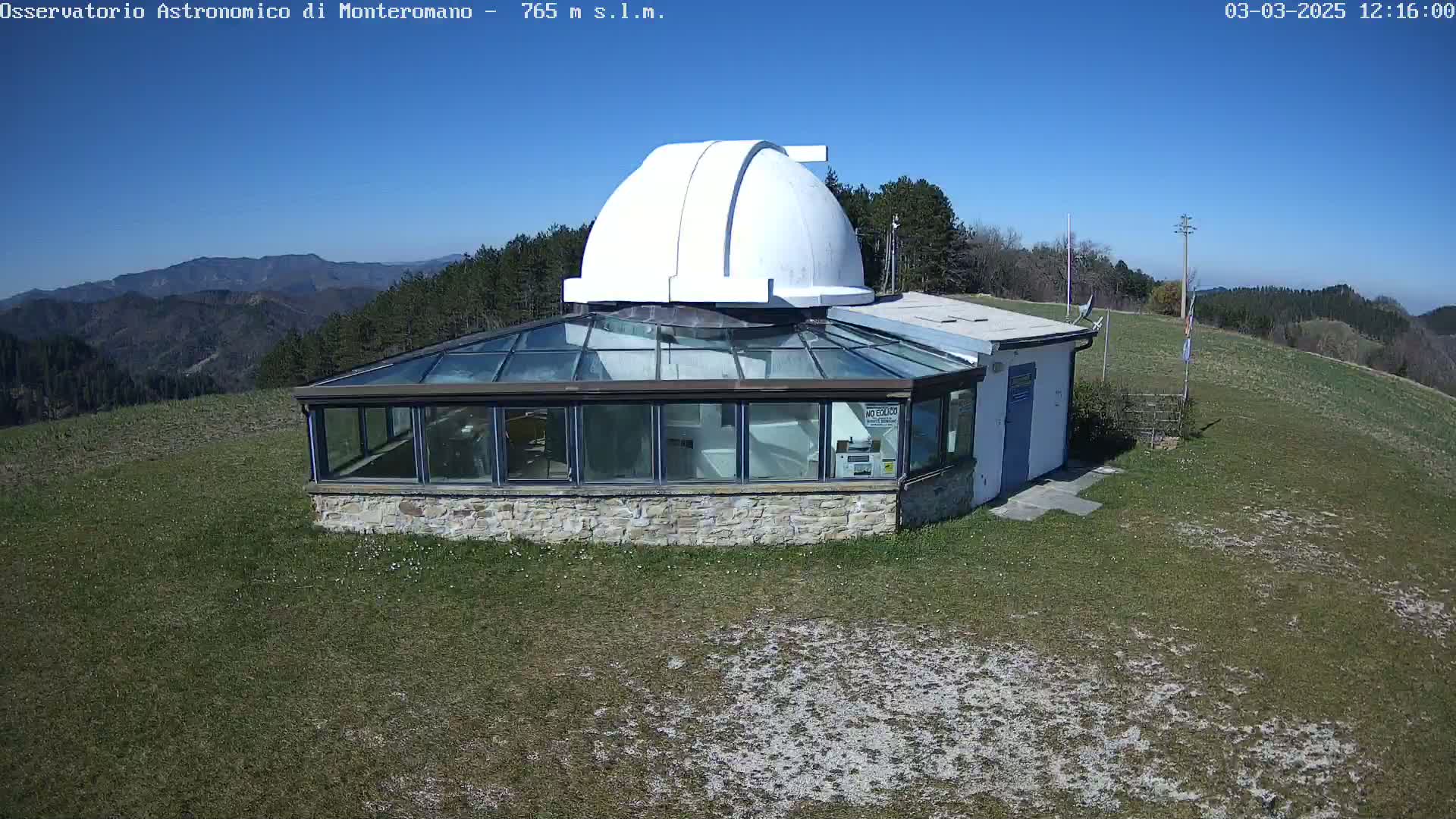 A white domed observatory building with a glass-roofed addition sits on a grassy hilltop under a clear blue sky.