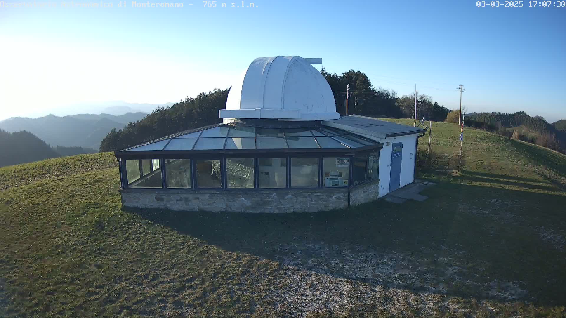 A white observatory dome sits atop a stone building with a glass sunroom, overlooking a grassy hillside and distant mountains under a clear blue sky.