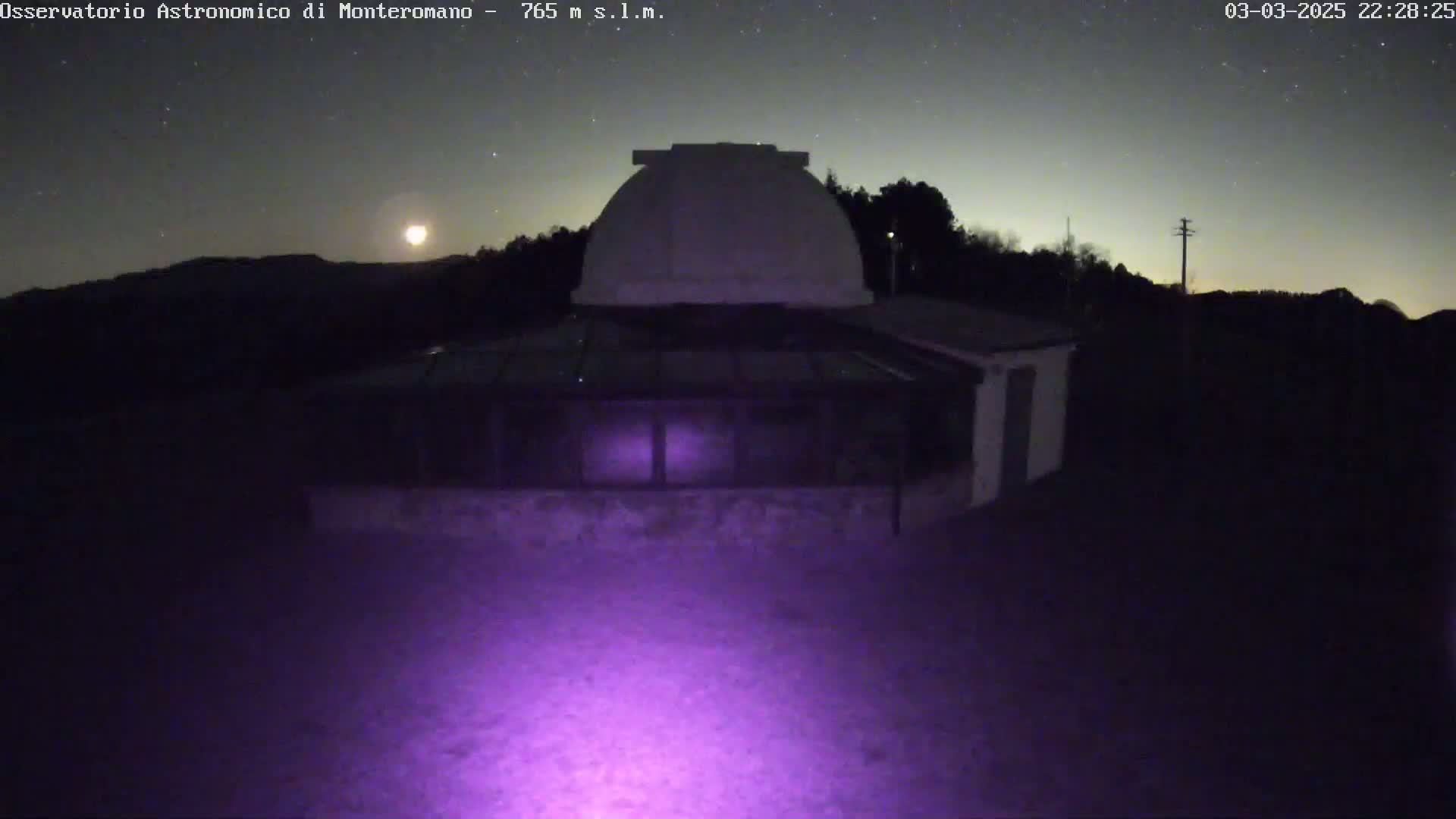Under a starry, moonlit night sky, a low-lying observatory building with a dome sits in the foreground, illuminated by a purple light.