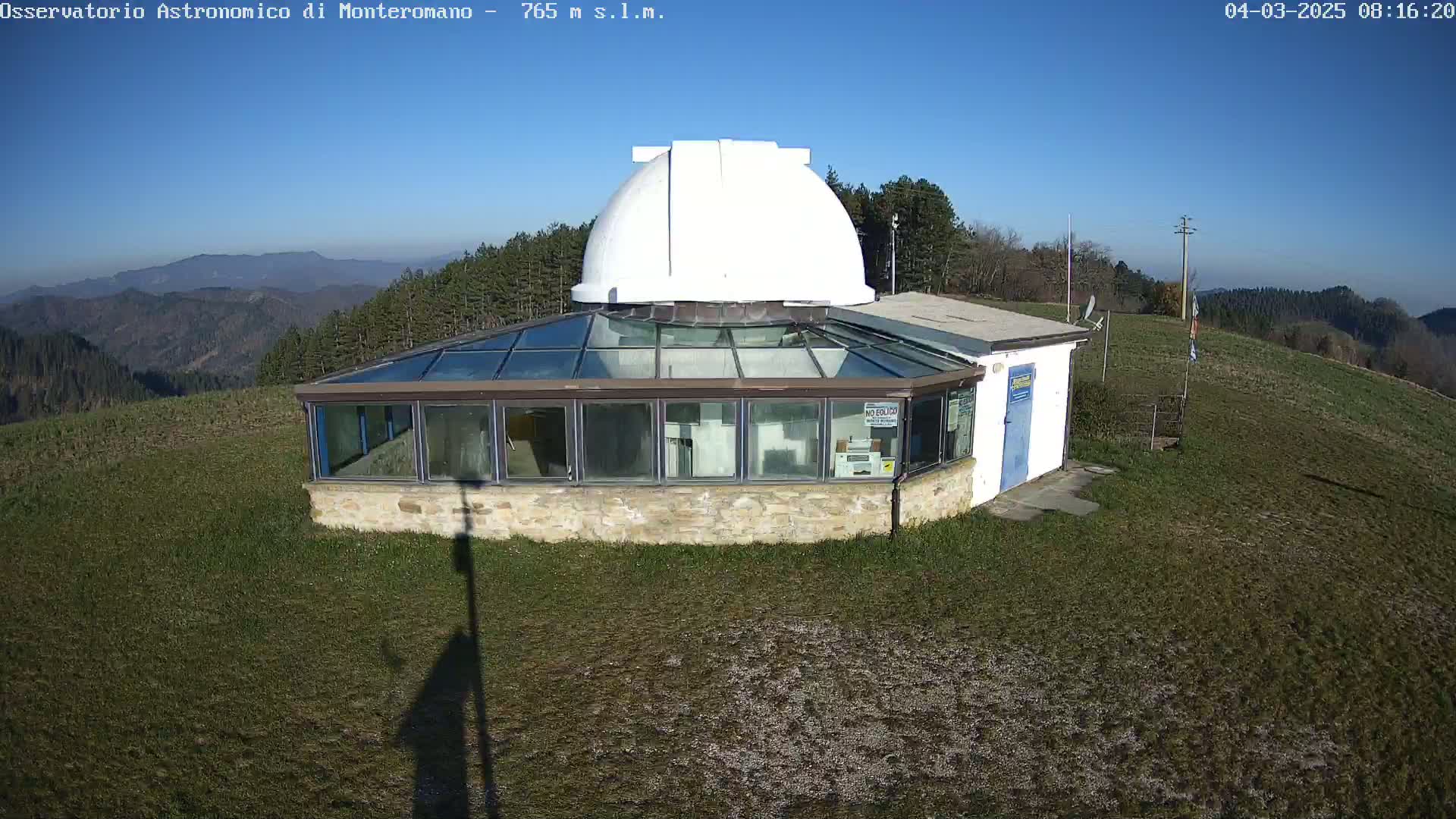 A white-domed observatory building with a glass-roofed addition sits atop a grassy hill under a clear blue sky.