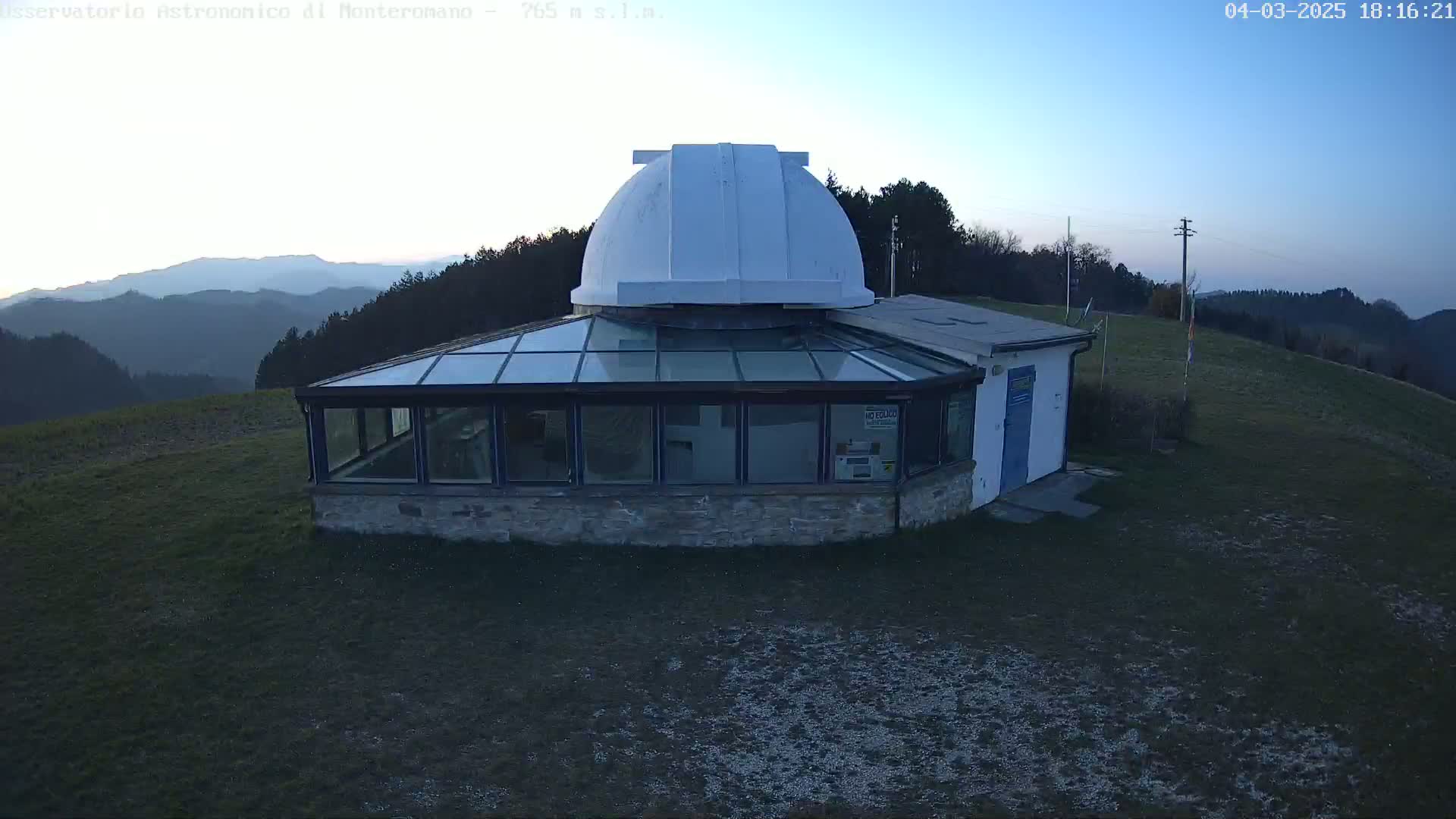 An astronomical observatory with a white dome sits atop a hill overlooking a mountain range under a twilight sky.