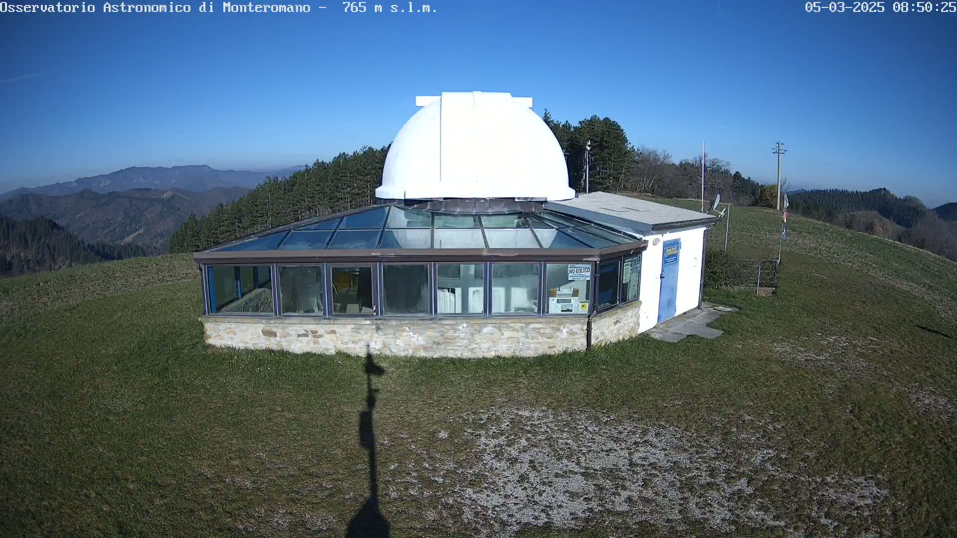 A white-domed observatory building with a glass-roofed addition sits on a grassy hilltop overlooking a mountain range under a clear blue sky.