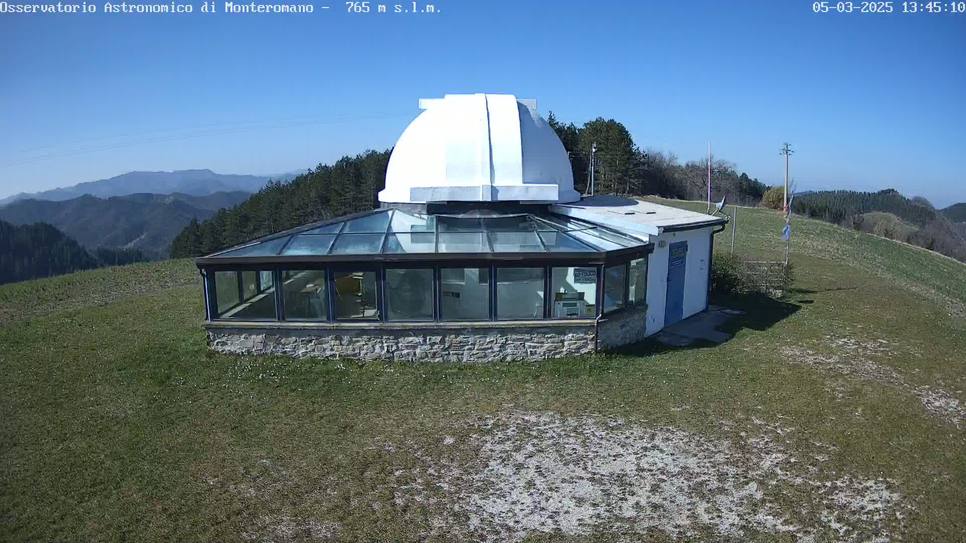 A stone building with a glass-roofed sunroom and a white dome sits on a grassy hilltop under a clear blue sky.