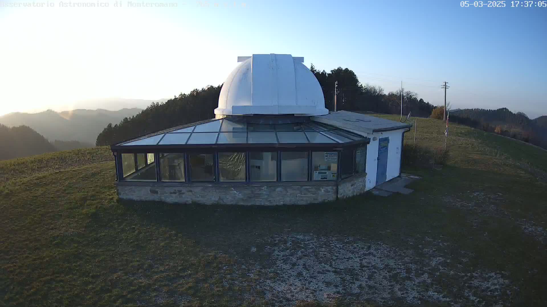 A white observatory dome sits atop a stone building with a glass-roofed addition, overlooking a grassy hill and distant mountains under a clear twilight sky.