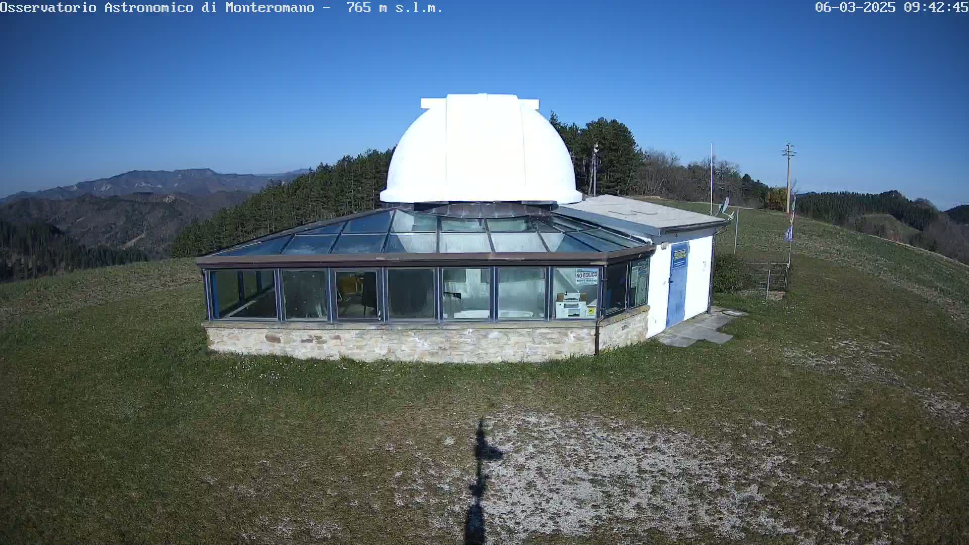 An observatory with a white dome sits atop a grassy hill on a clear, sunny day, overlooking a distant mountain range.
