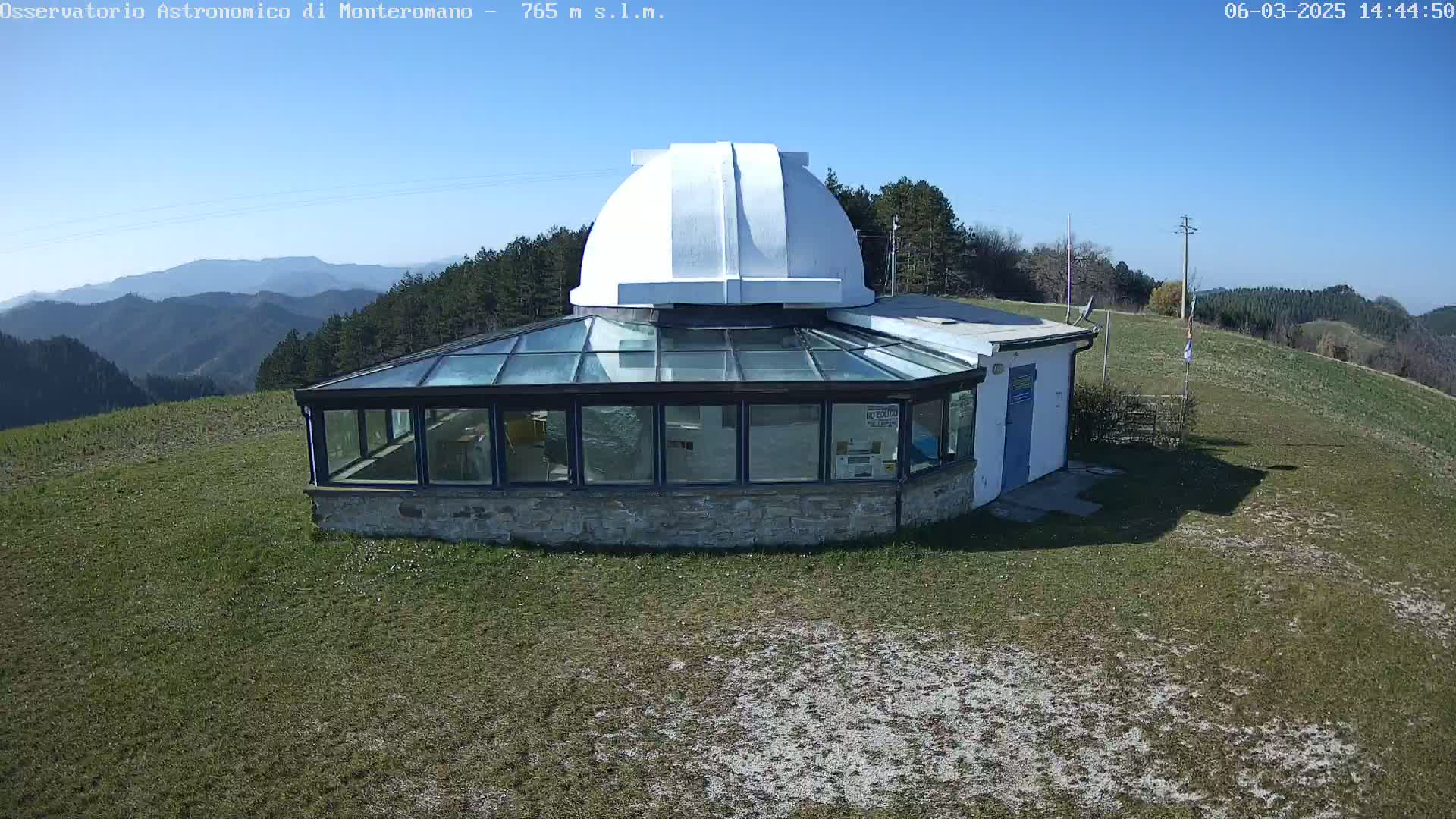 A glass-roofed observatory building with a white dome sits on a grassy hilltop overlooking a mountain range under a clear blue sky.