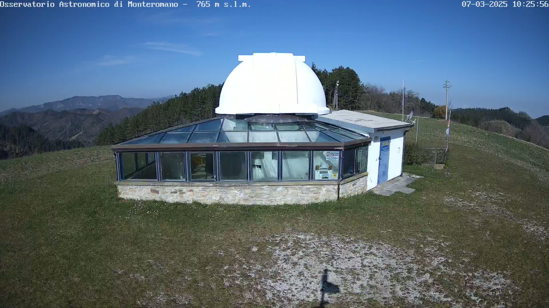 A white dome observatory building with a glass-roofed addition sits on a grassy hilltop under a clear blue sky.