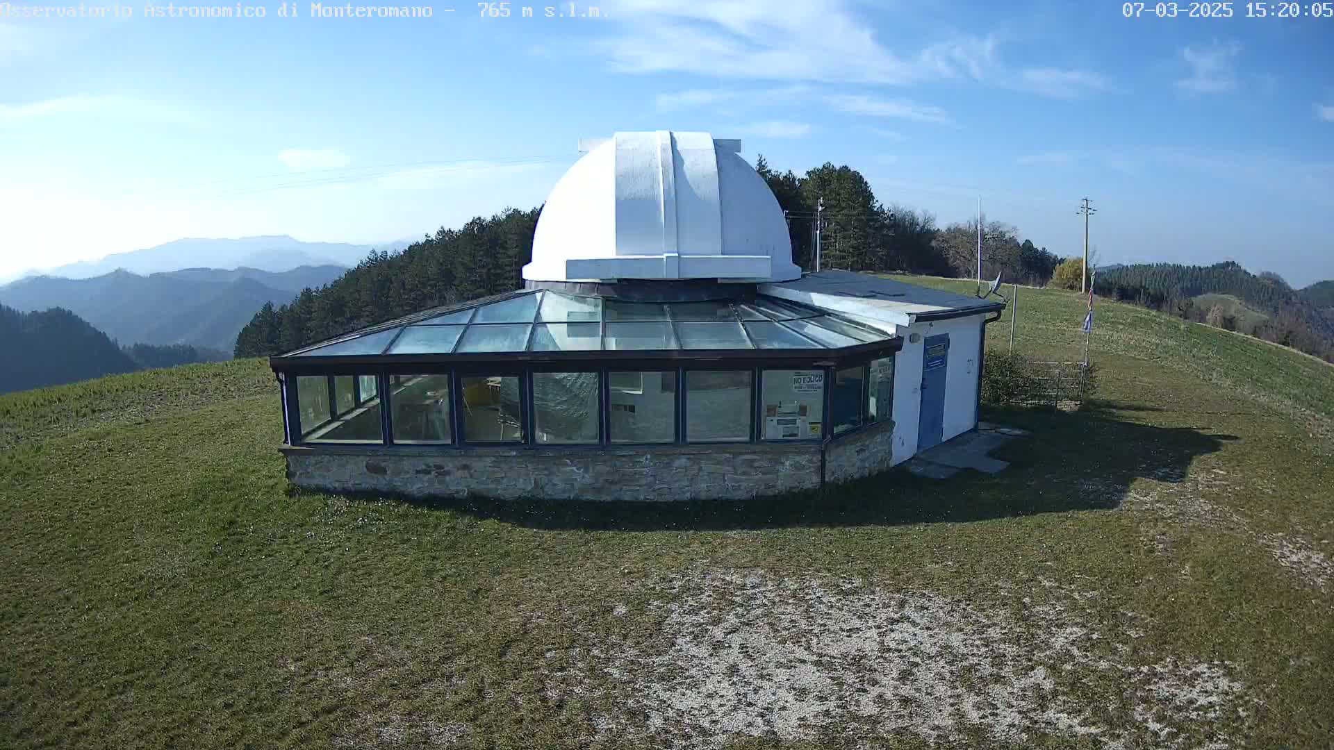 A white domed observatory building with a glass sunroom sits on a grassy hilltop under a mostly sunny sky, overlooking a distant mountain range.