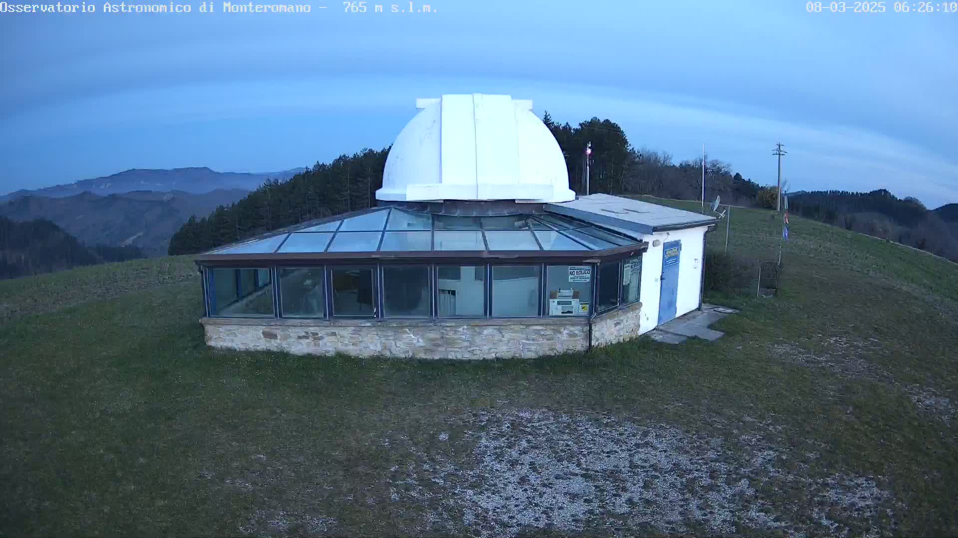 A white-domed observatory building with a glass-roofed addition sits on a grassy hilltop overlooking a range of distant, hazy mountains under a twilight sky.