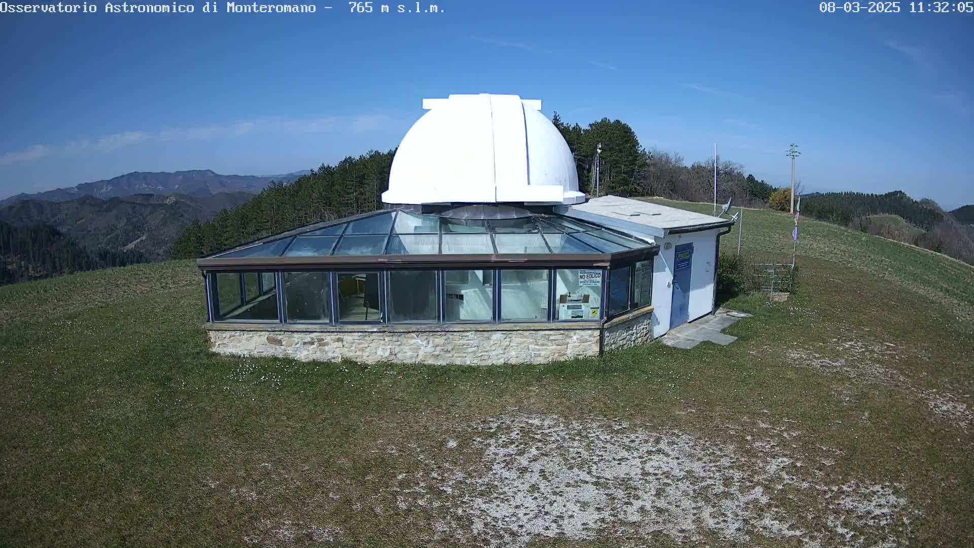 A stone building with a glass roof and a white dome on top sits on a grassy hill overlooking a mountain range under a clear blue sky.