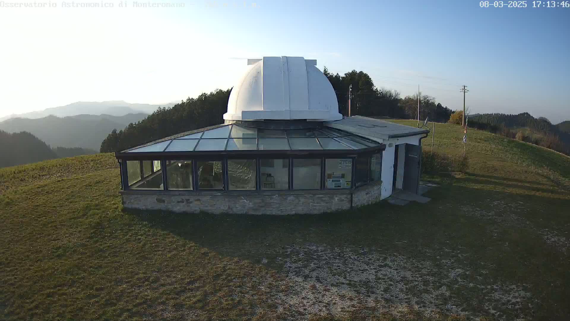 A stone building with a glass-roofed addition and a white dome sits on a grassy hilltop under a clear blue sky.