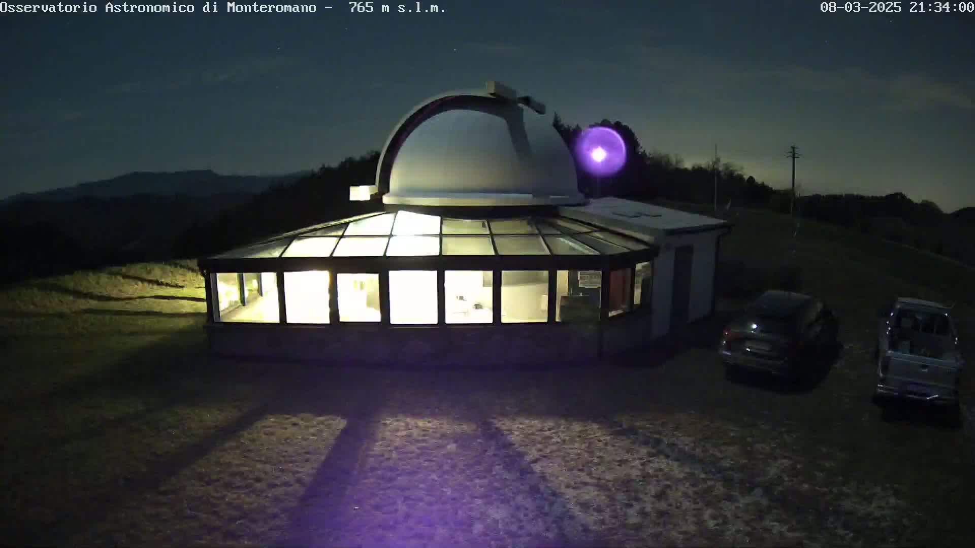 An illuminated observatory building with a dome, a glass-roofed addition, and two cars parked outside, all under a dark, clear night sky.