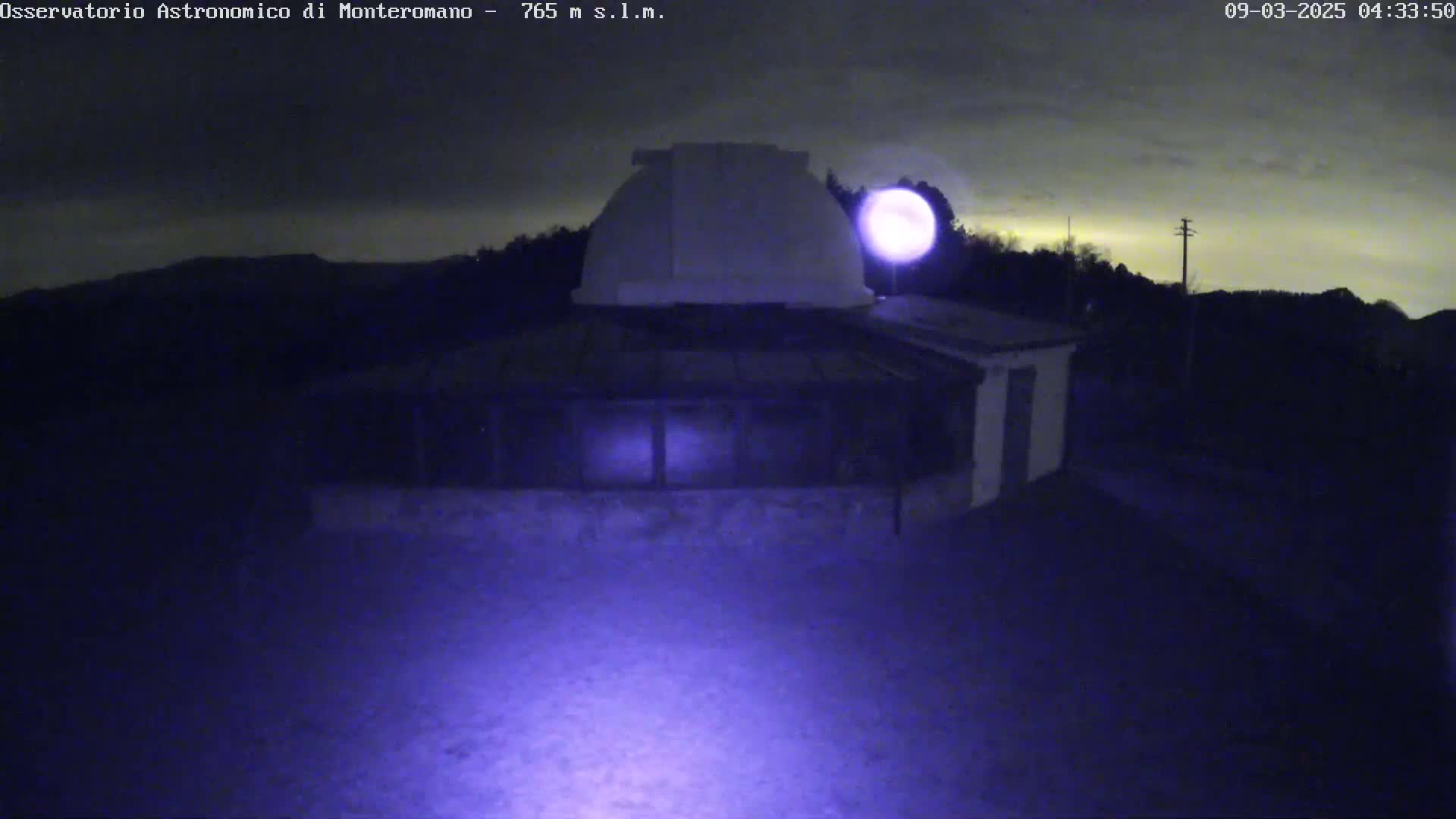 A low-light nighttime image shows a building with a dome-shaped roof, possibly an observatory, under a mostly clear, dark sky with a bright moon or light source visible.