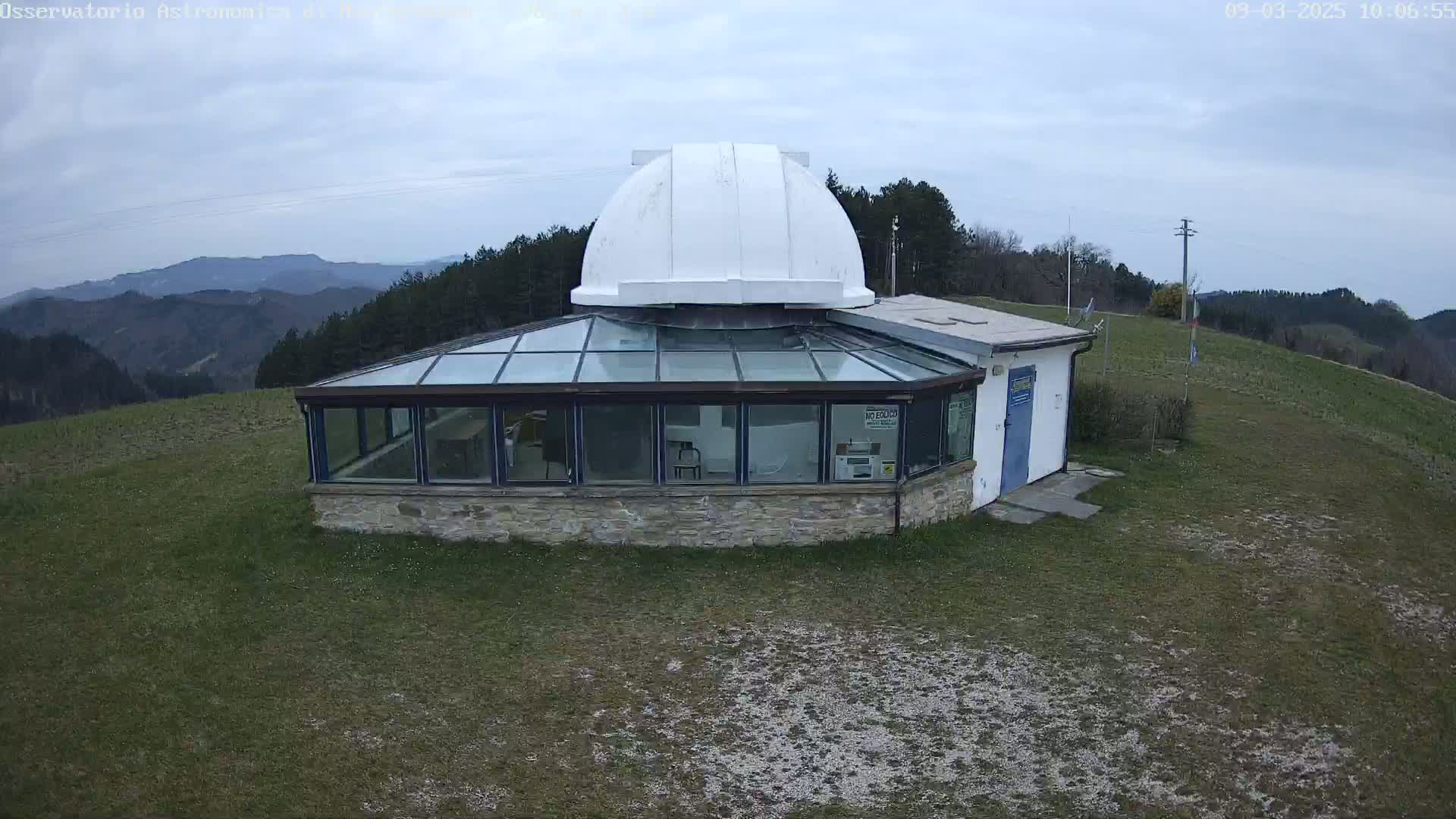 An observatory with a white dome sits atop a grassy hill overlooking a mountainous landscape under a mostly cloudy sky.