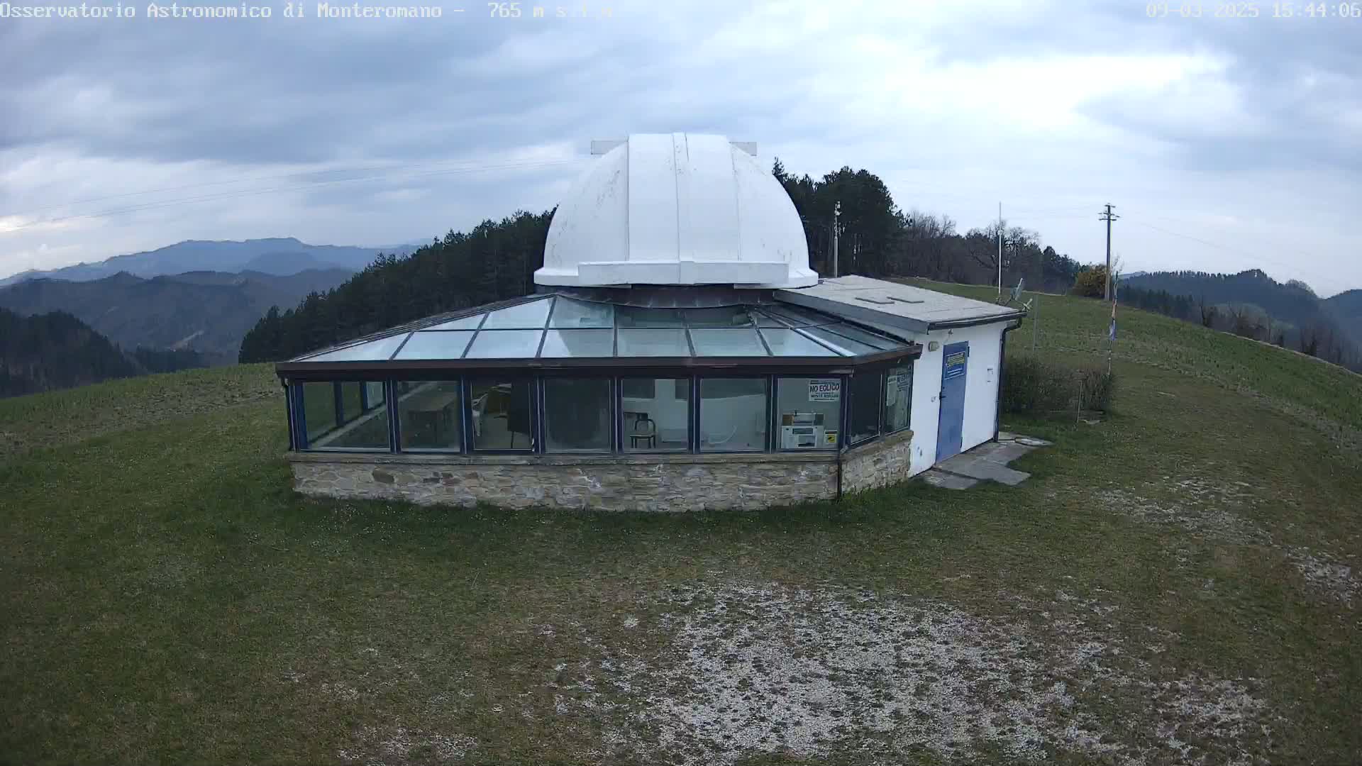 A white dome observatory sits atop a grassy hill overlooking a mountain range under a cloudy sky.