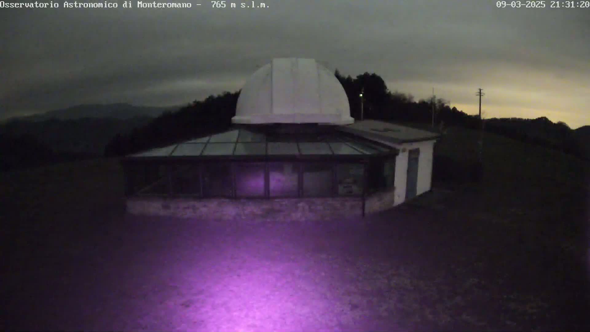 A white dome-shaped observatory building with a glass-roofed addition is illuminated by a purple light at night under a dark, cloudy sky.