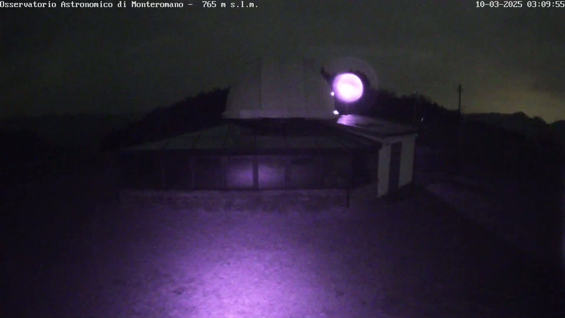 A nighttime, low-light image shows a building with a dome-shaped roof and a glass-roofed addition, illuminated by a purple light source.