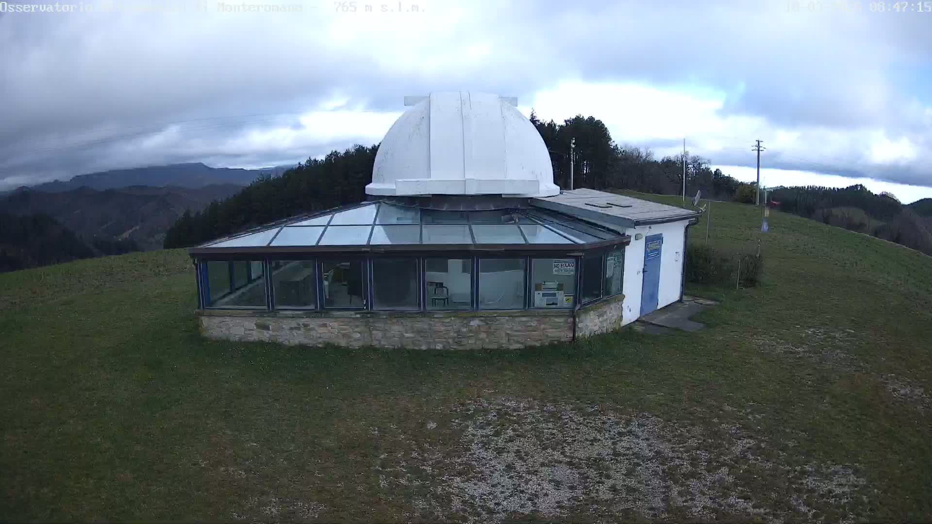 An observatory with a white dome sits atop a grassy hill under a mostly cloudy sky.