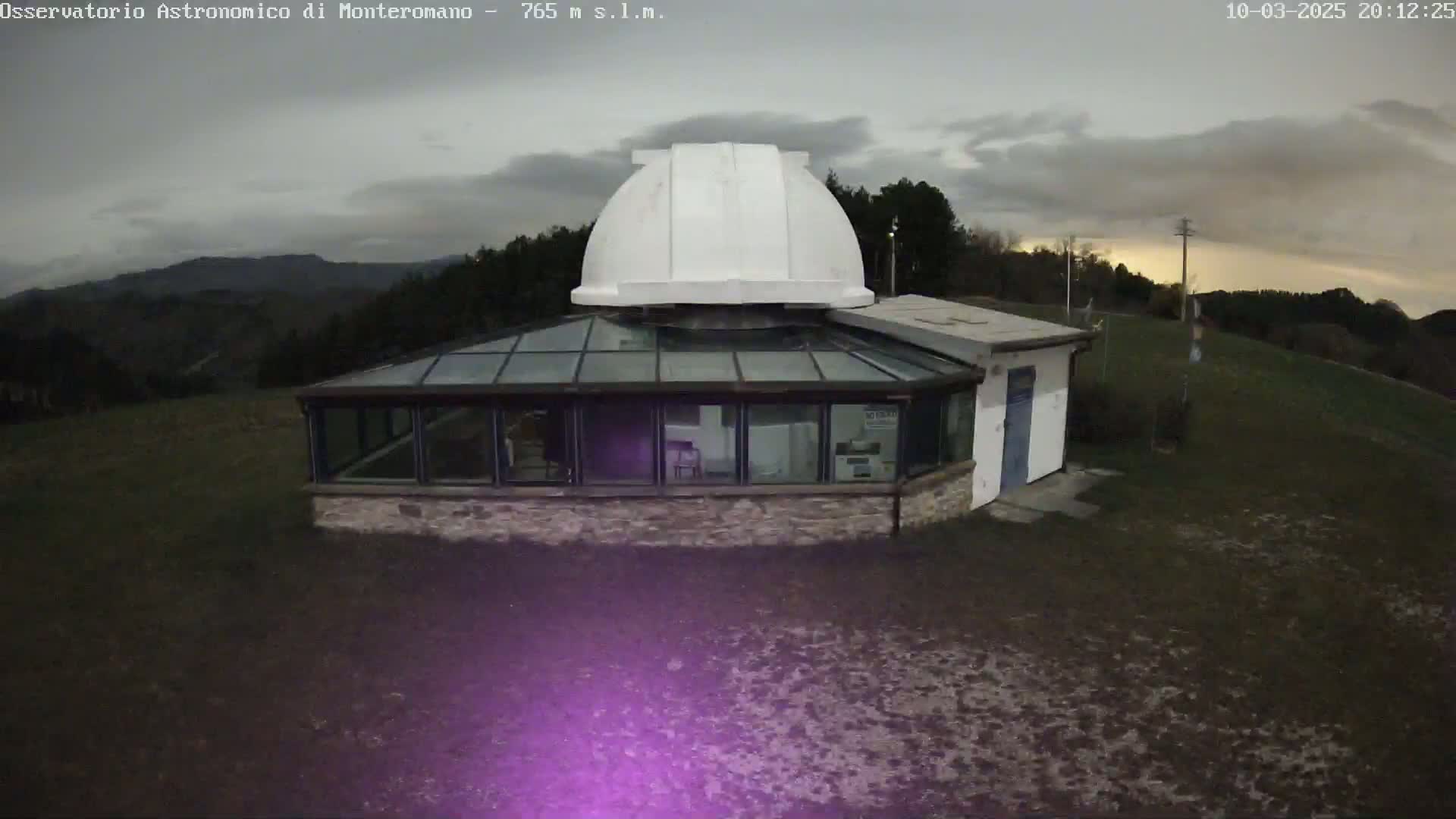 A white observatory dome sits atop a stone building with a glass sunroom, situated on a hill under an overcast sky.