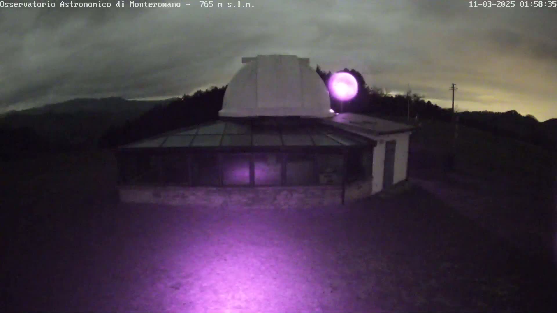 An observatory building with a dome is illuminated by a purple light under a cloudy night sky.