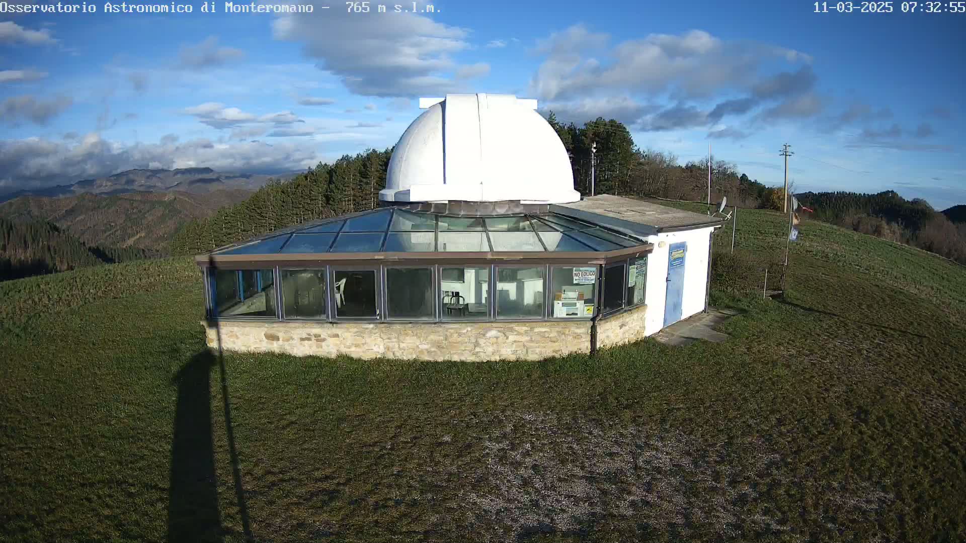 An observatory with a white dome and a glass-roofed building sits on a grassy hilltop under a partly cloudy blue sky.