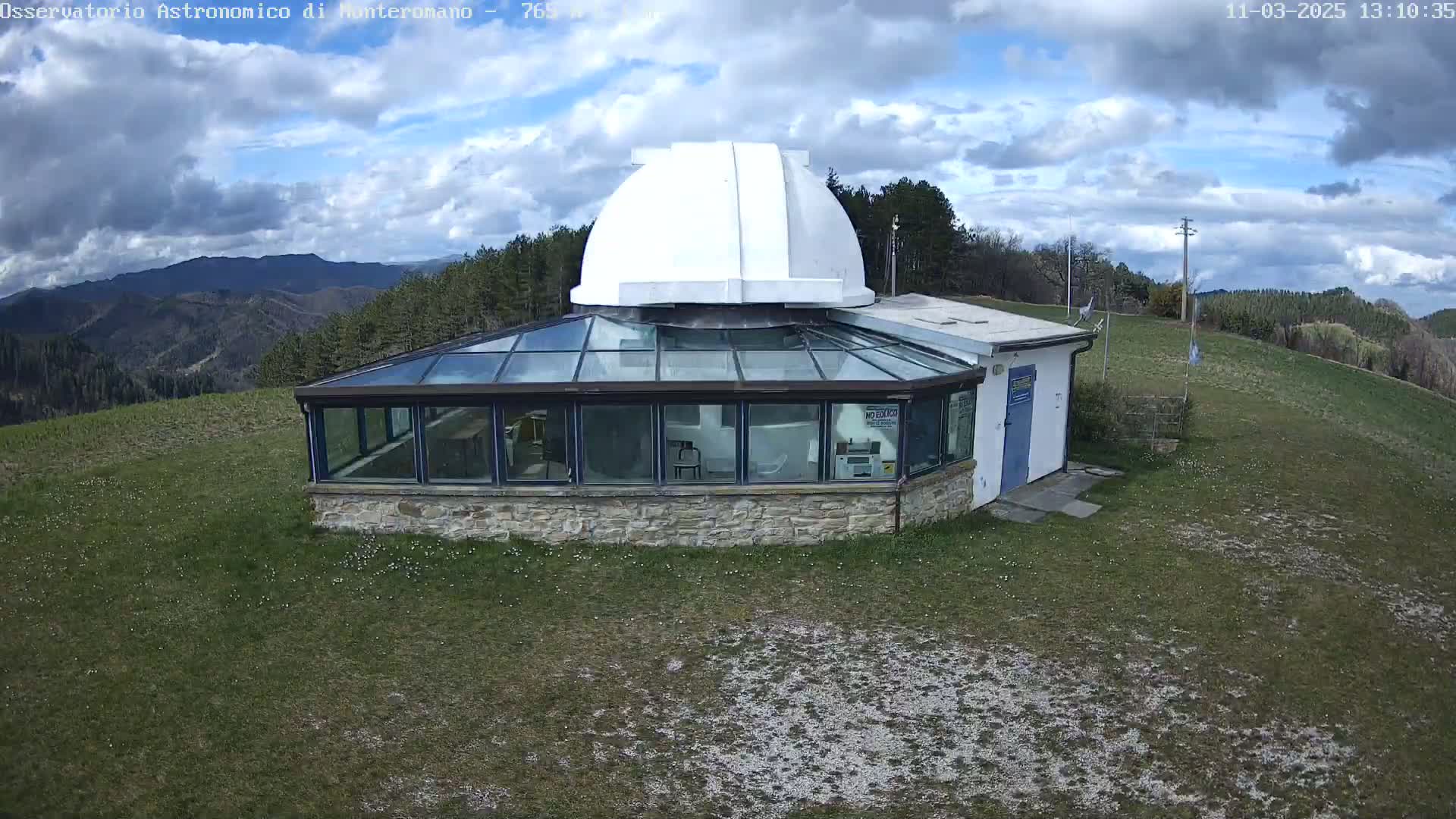 A stone building with a glass roof and a white dome sits on a grassy hill overlooking a mountain range under a partly cloudy sky.