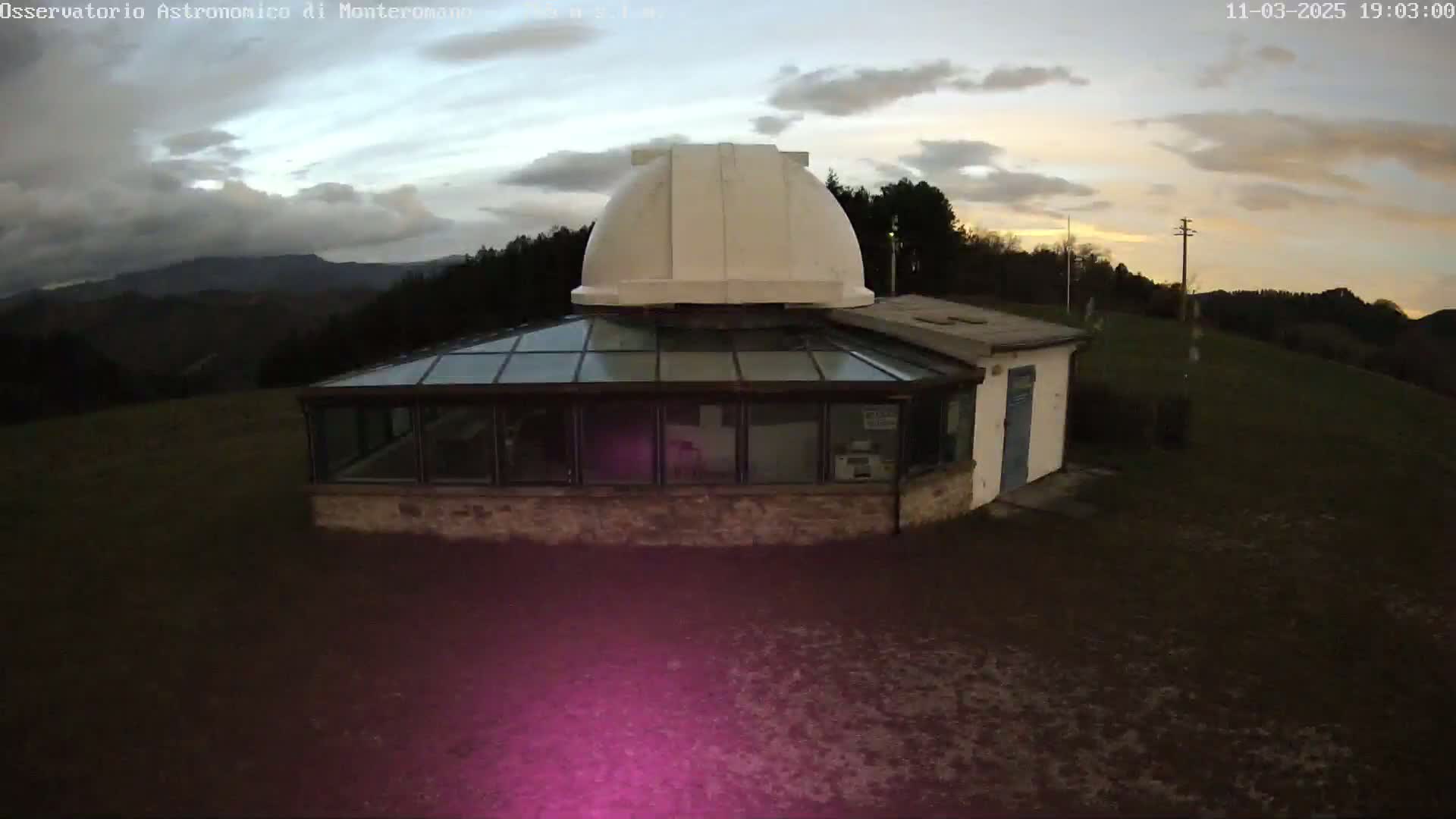 A small stone building with a glass conservatory and a white dome sits on a grassy hill under a partly cloudy twilight sky.