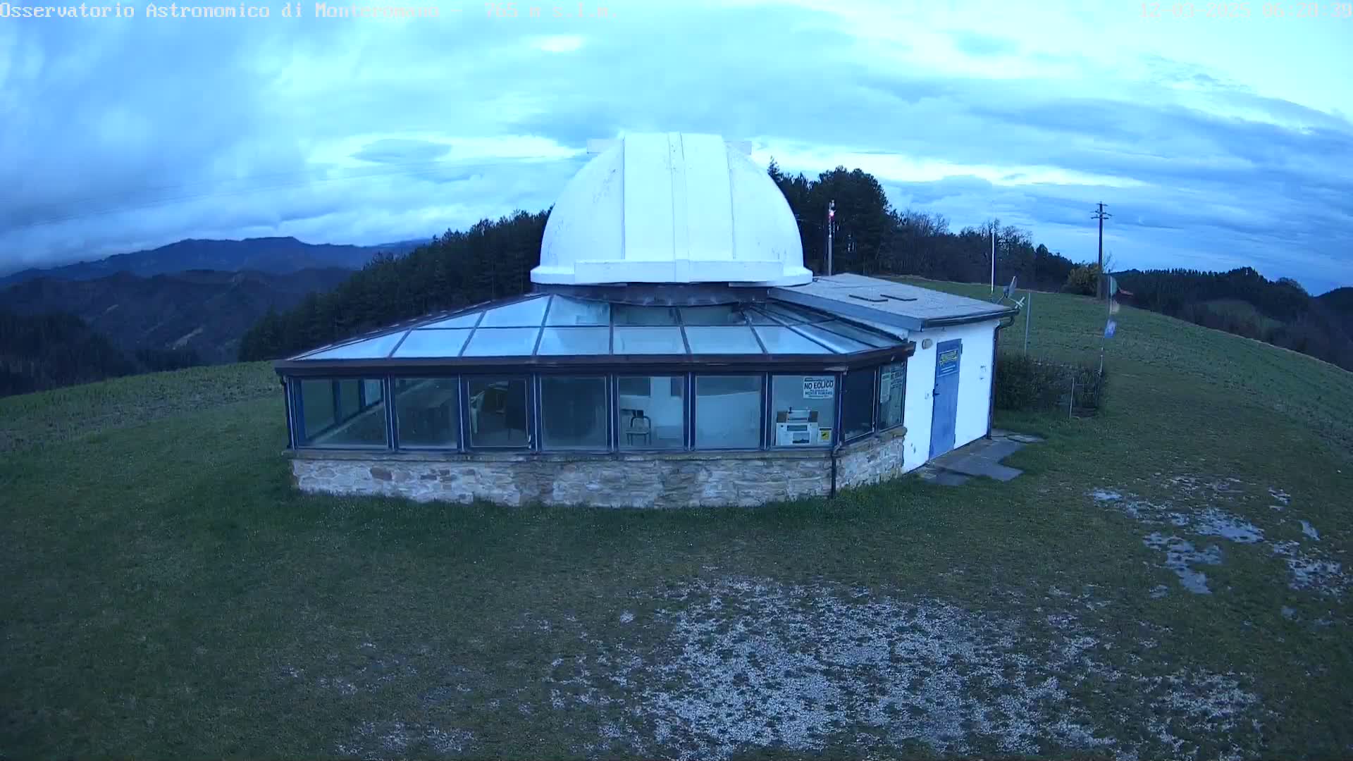 A stone building with a glass-roofed addition and a white dome sits on a grassy hilltop under a cloudy sky.