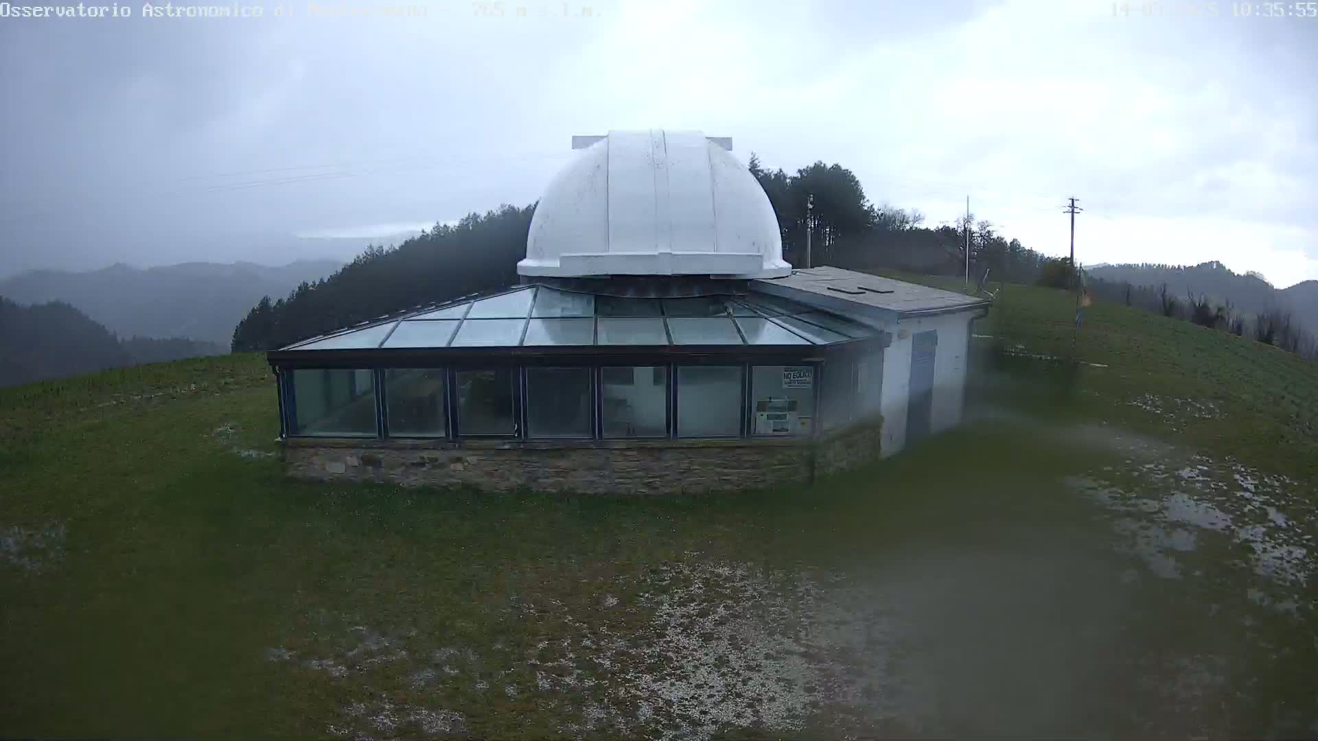 A white-domed observatory building with a glass-roofed addition sits on a grassy hill overlooking a misty mountain range under an overcast sky.