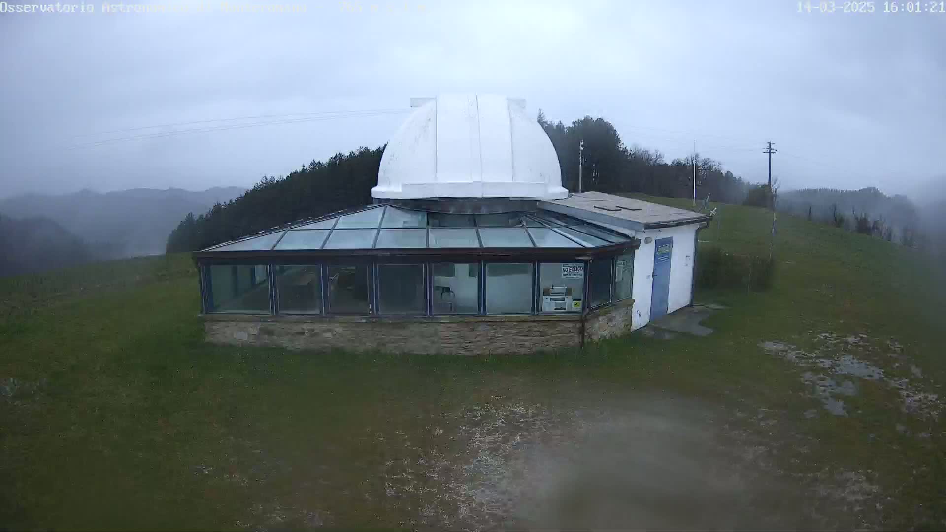 A white-domed observatory building with a glass-roofed addition sits on a grassy hill overlooking a misty, overcast landscape.