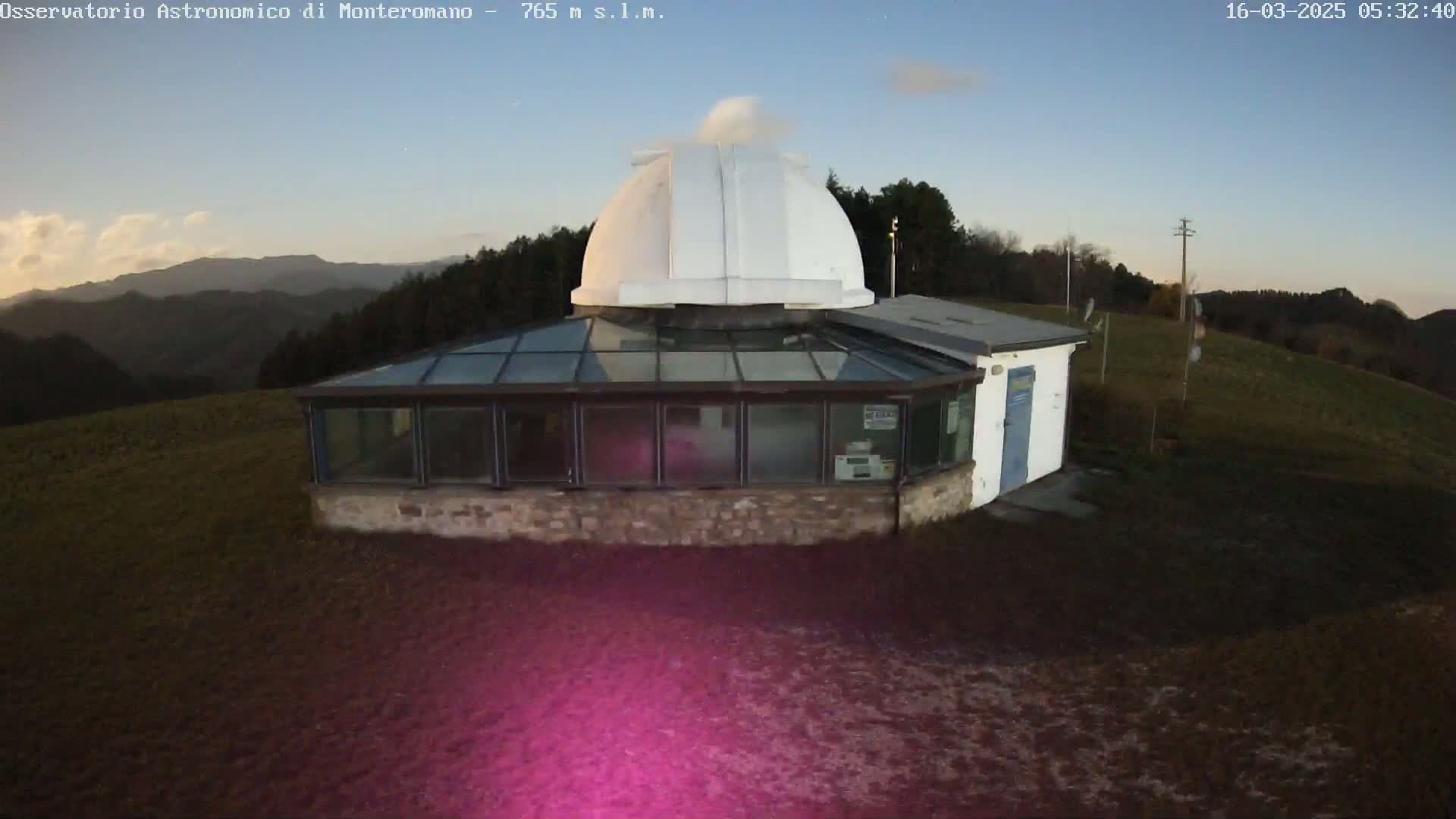 An astronomical observatory with a white dome and glass-roofed addition sits on a grassy hilltop under a twilight sky.
